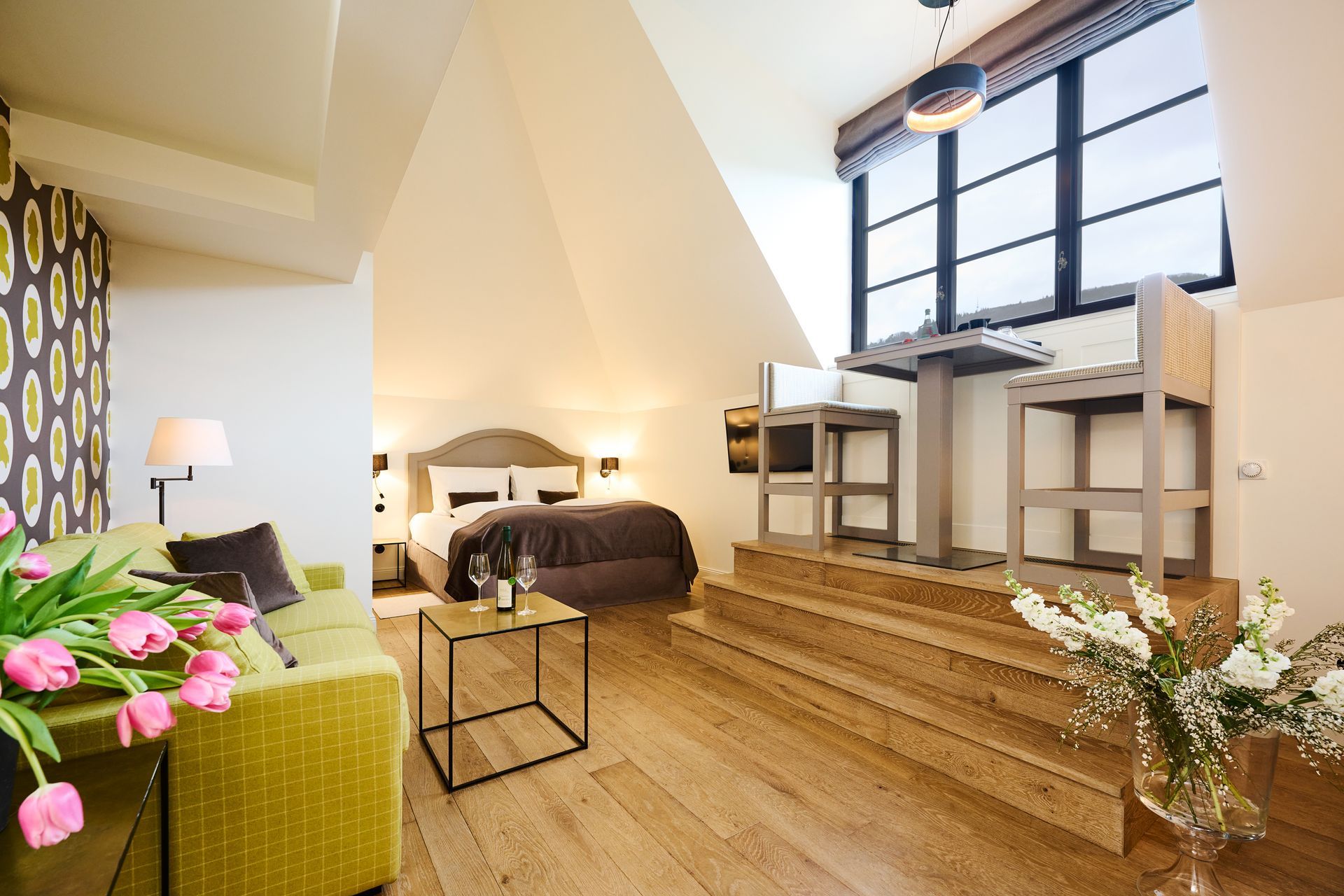 Ein gemütliches Dachgeschoss-Hotelzimmer mit einem grünen Sofa, einem Bett und einem erhöhten Essbereich mit zwei Stühlen an einem großen Fenster.