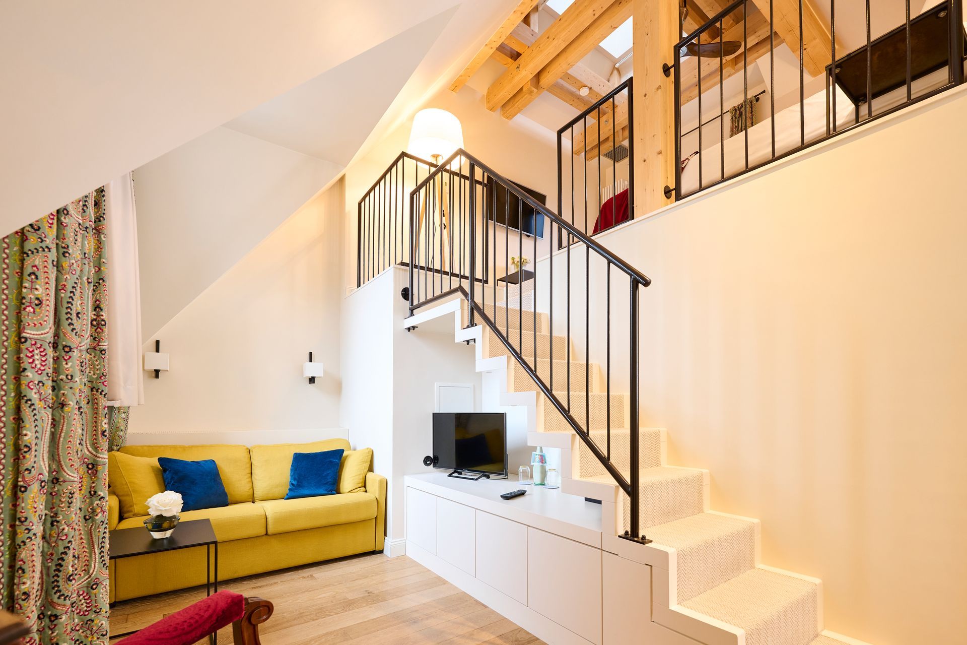 In einem gemütlichen Zimmer mit einer Treppe, die zu einem Dachboden führt, steht ein gelbes Sofa mit blauen Kissen.