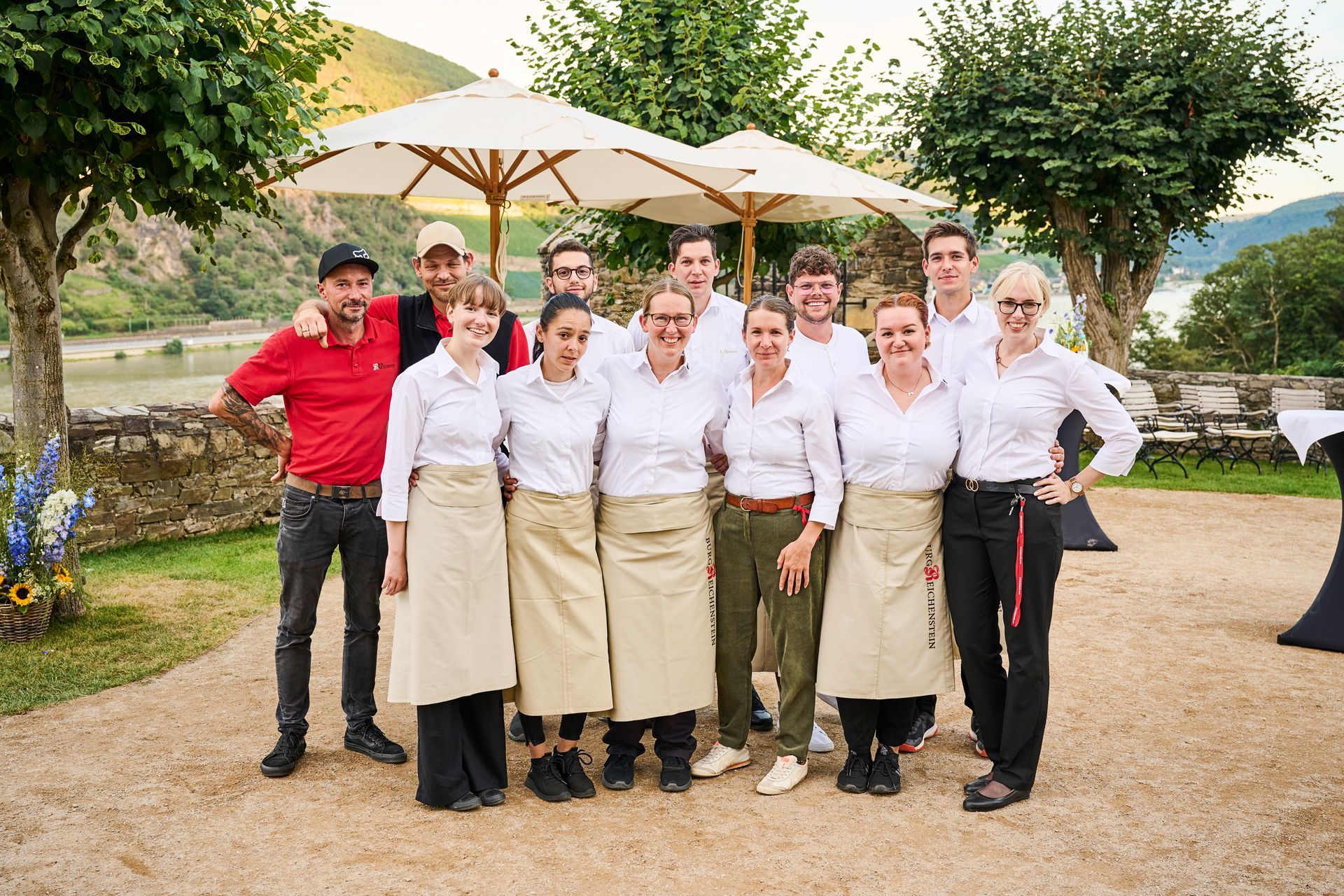 Eine Gruppe von Mitarbeitern in weißen Hemden und beigen Schürzen steht lächelnd unter Sonnenschirmen im Freien an einem Fluss.