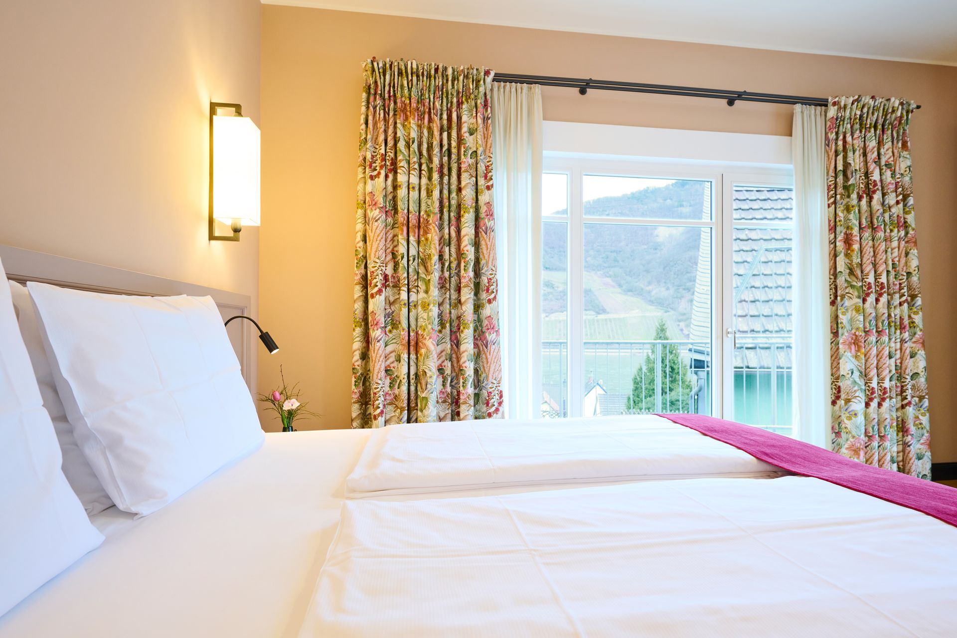 Ein sauberes Hotelbett mit weißer Bettwäsche und einer magentafarbenen Tagesdecke, neben einem großen Fenster mit geblümten Vorhängen und Bergblick.