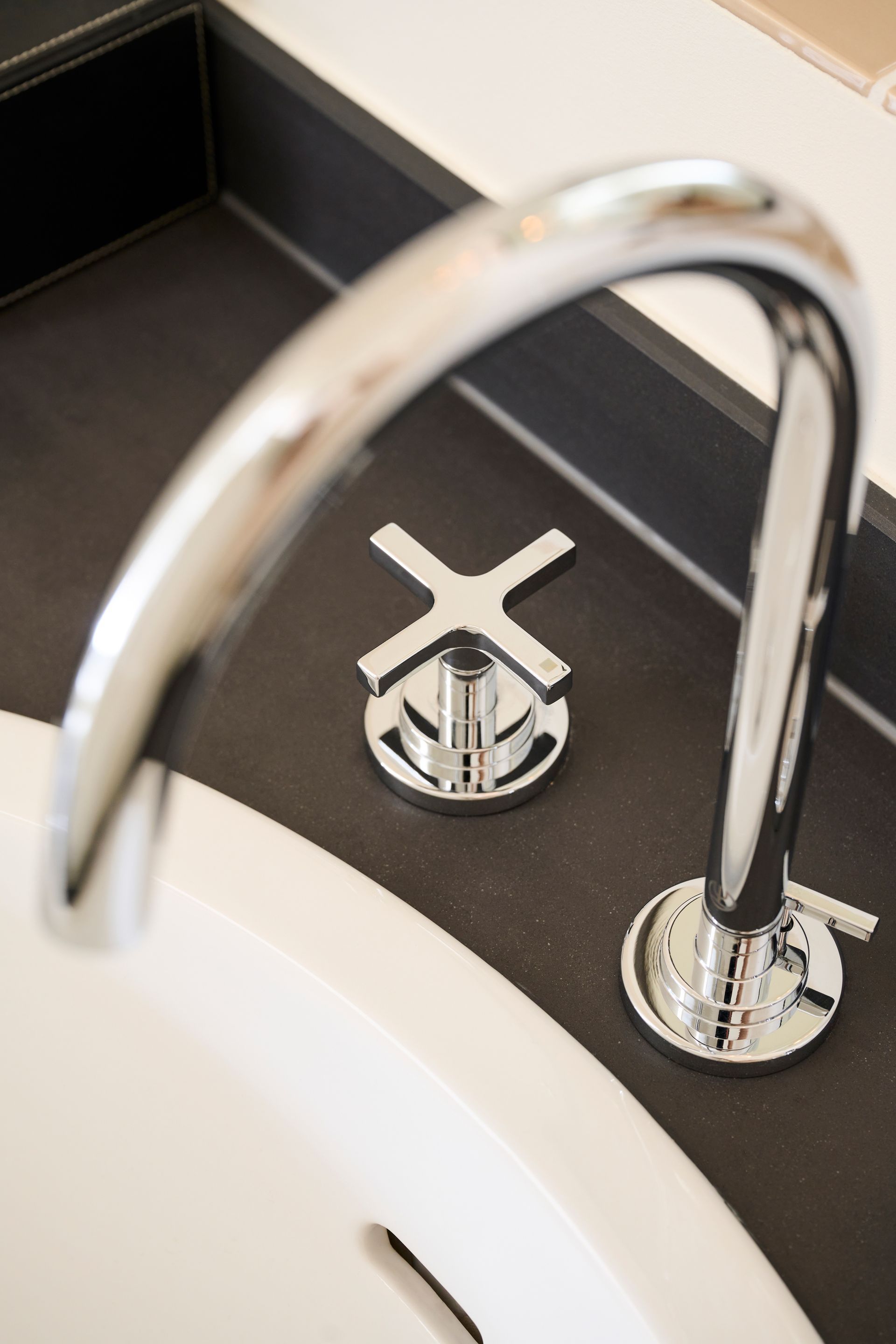 Ein polierter Chrom-Wasserhahn mit Kreuzgriffsteuerung ist auf einer dunklen, matten Arbeitsplatte über einem weißen Waschbecken montiert.