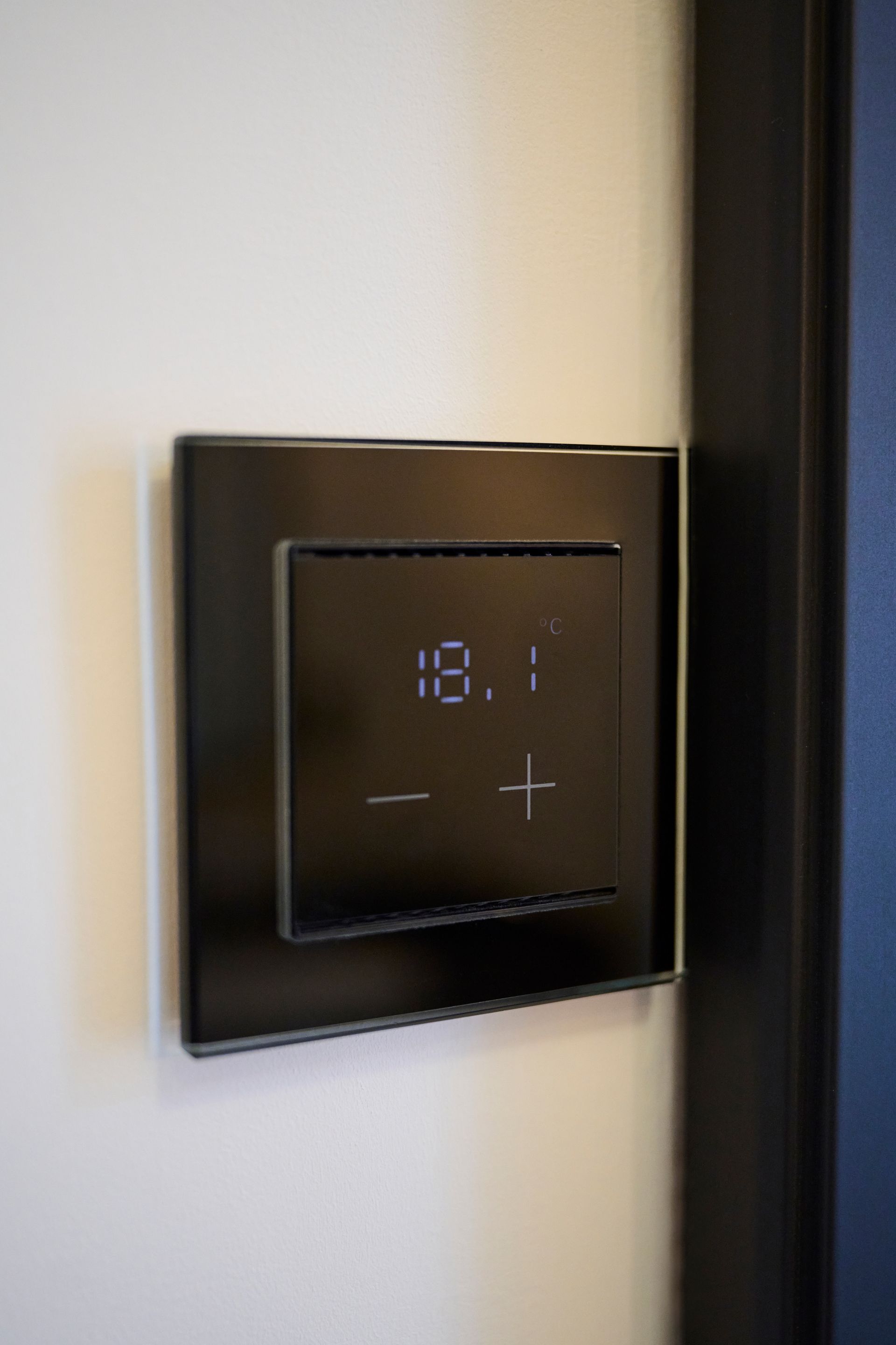Ein schwarzer digitaler Thermostat, der an einer weißen Wand montiert ist und eine Temperatur von 18,1 anzeigt, mit Plus- und Minus-Touch-Bedienelementen.