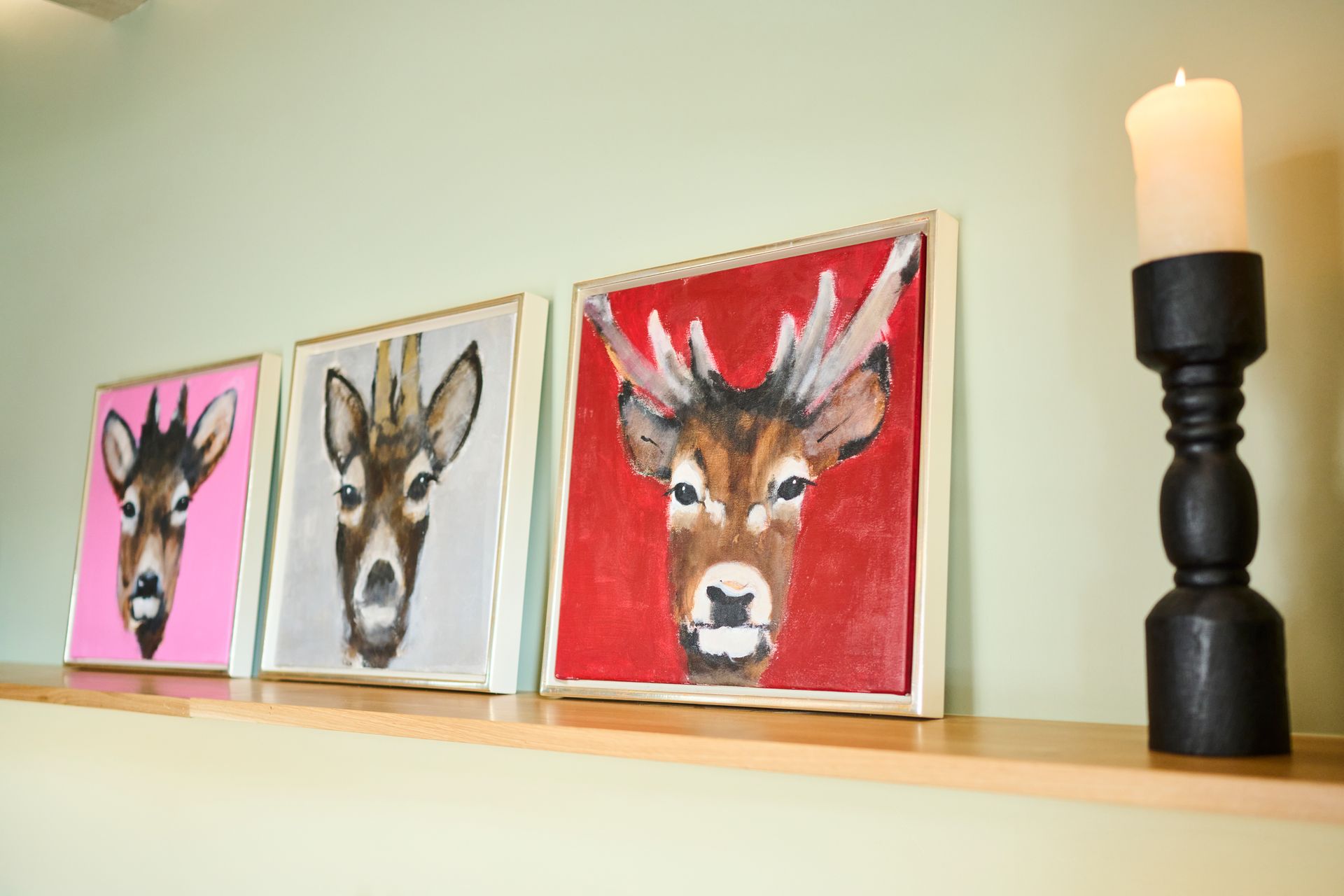 Drei quadratische Gemälde von Hirschköpfen in rosa, grauen und roten Rahmen stehen auf einem Regal neben einem schwarzen Kerzenhalter.