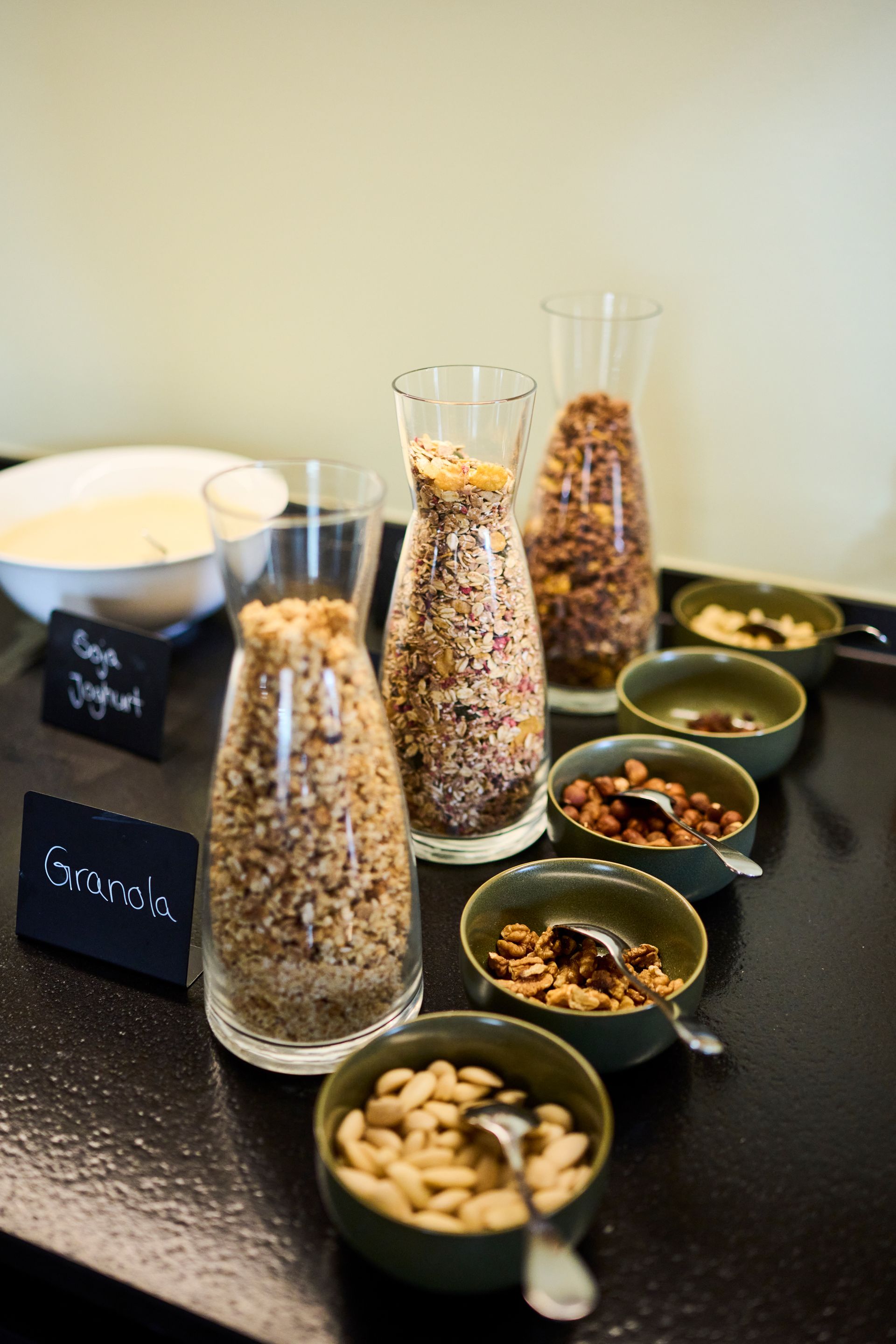 Ein Frühstücksbuffet mit durchsichtigen Karaffen mit Müsli und Schüsseln mit verschiedenen Nüssen, Samen und Joghurt auf einer dunklen Oberfläche.