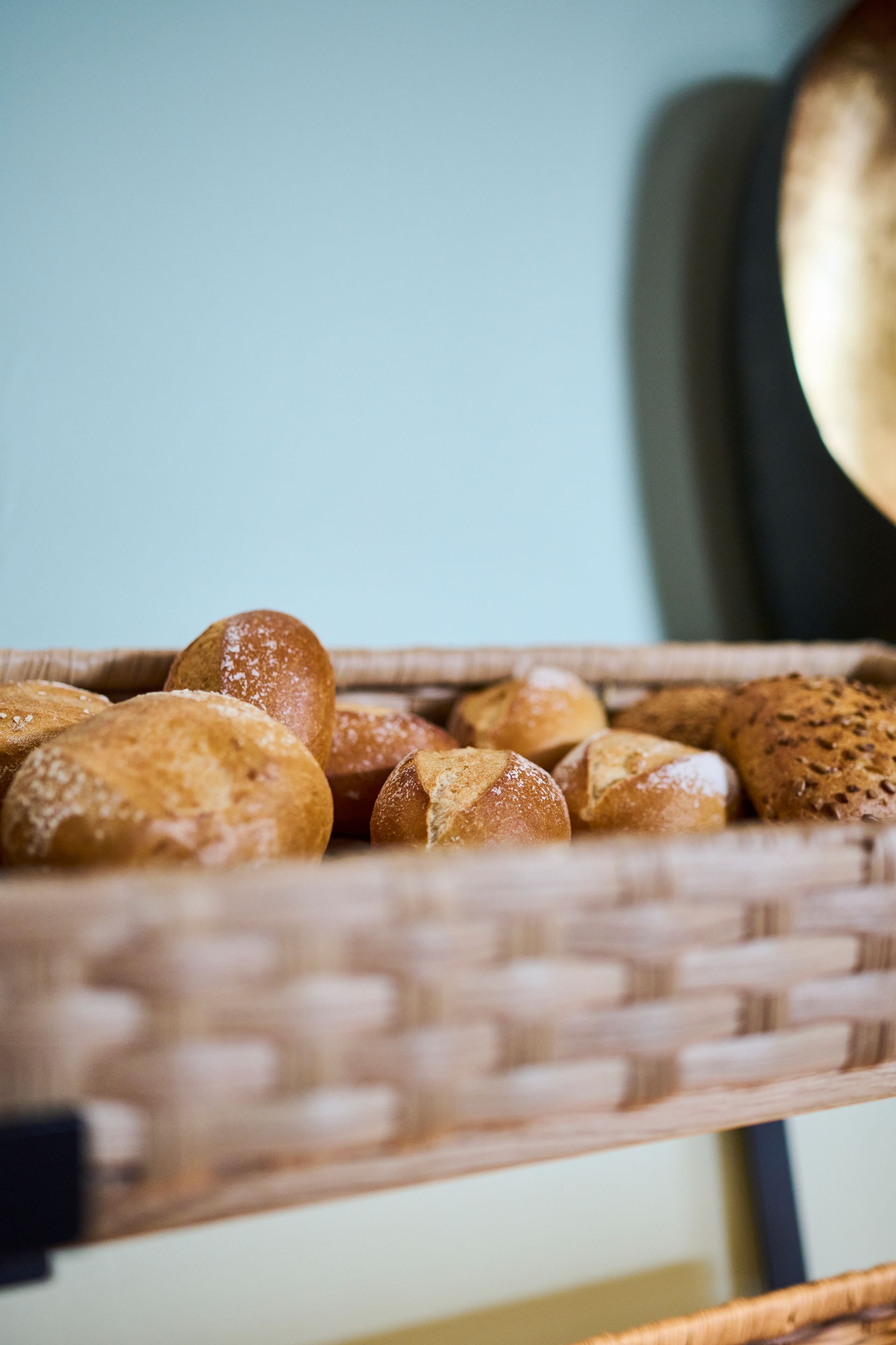 Ein geflochtener Korb, gefüllt mit verschiedenen goldbraunen Brötchen, vor einer verschwommenen türkisfarbenen Wand und einem dunklen Dekorationsobjekt.