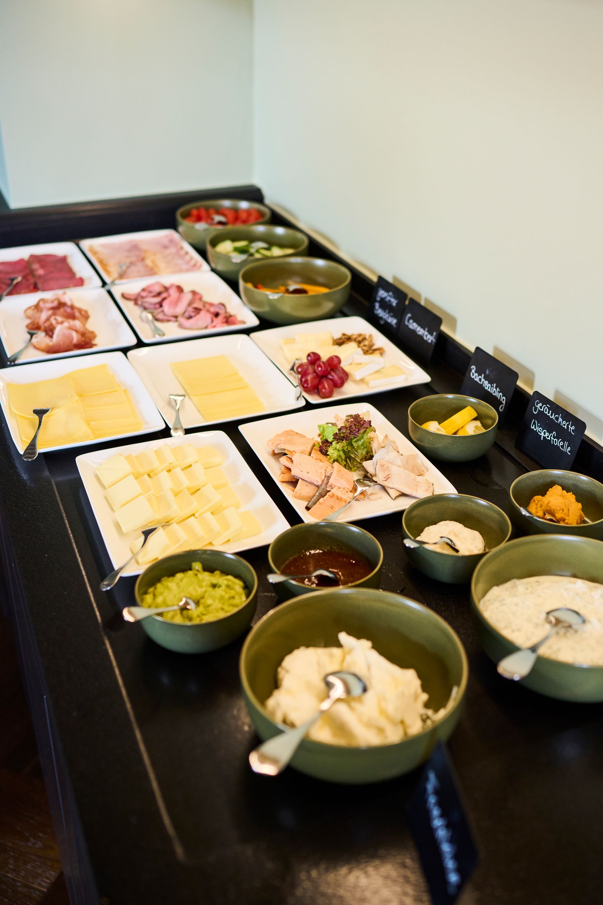 Ein reichhaltiges Frühstücksbuffet mit verschiedenen Fleischsorten, Käse, Früchten und Schüsseln mit Toppings auf einer dunklen Oberfläche.