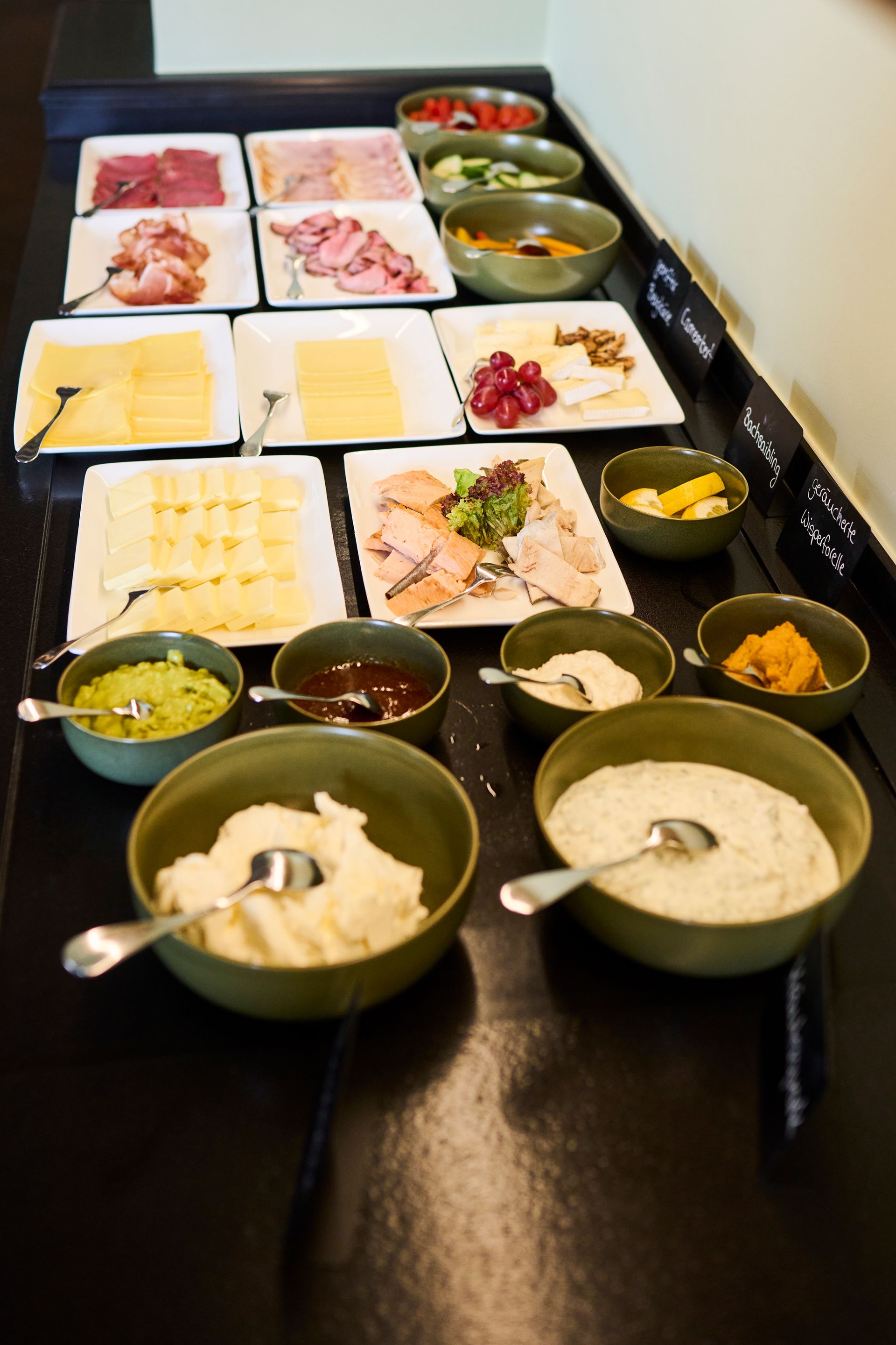 Eine Draufsicht auf ein Frühstücksbuffet mit verschiedenen Fleischsorten, Käse, Schüsseln mit Aufstrichen und frischem Obst.