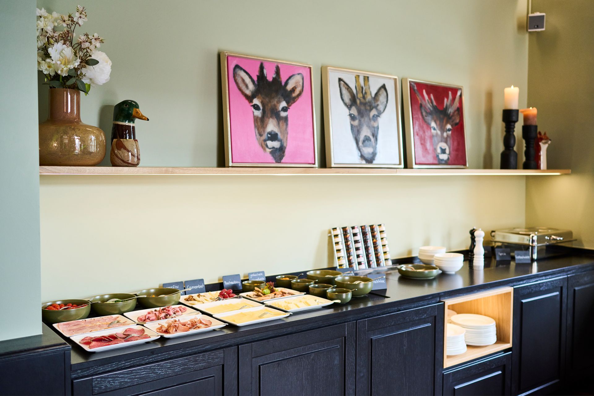 Ein Frühstücksbuffet mit dunklen Holzschränken, einer hellen Wand und drei Tierporträts, die darüber auf einem Regal hängen.