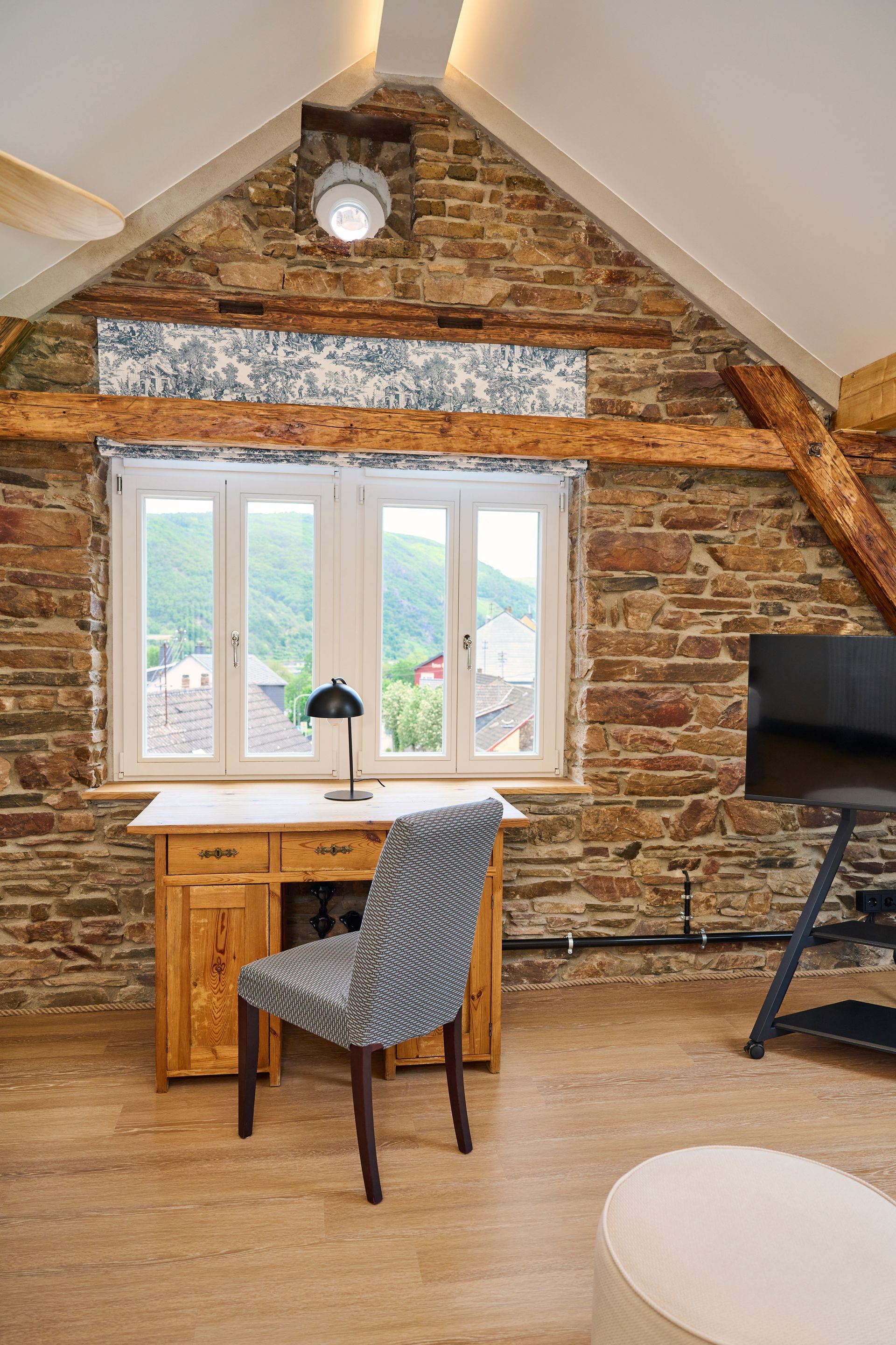 In einem Dachzimmer mit Dachschräge stehen vor einem Fenster an einer rustikalen Steinwand ein hölzerner Schreibtisch und ein gemusterter Stuhl.