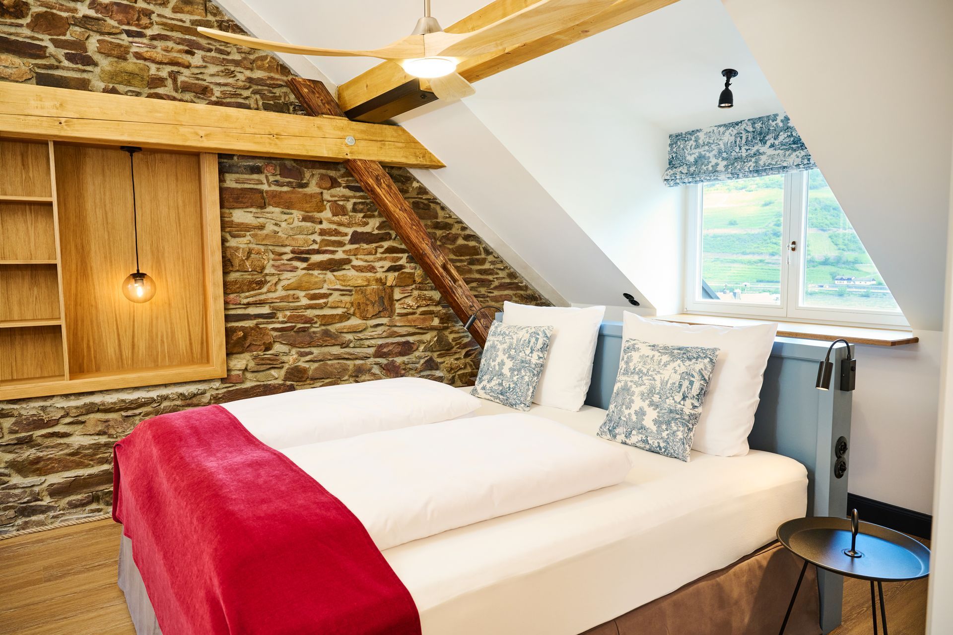 Ein rustikales Schlafzimmer mit Steinwänden, einem Holzbalken an der Decke, einem Bett mit weißer Bettwäsche, einer rosa Tagesdecke und einem Fenster mit Aussicht.