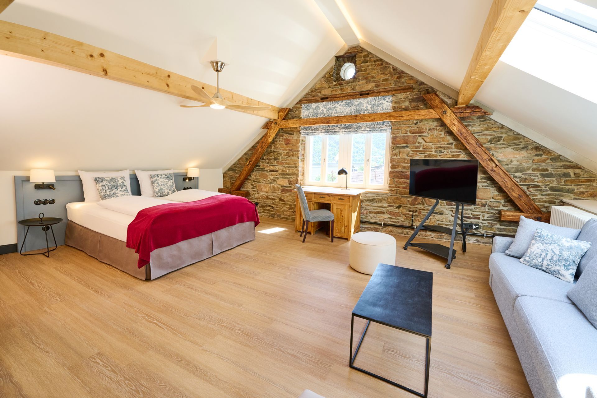 Ein Schlafzimmer im Dachgeschoss mit freiliegenden Holzbalken, Steinwänden, einem Bett mit roter Tagesdecke, einem Schreibtisch, einem Fernseher und einem grauen Sofa.