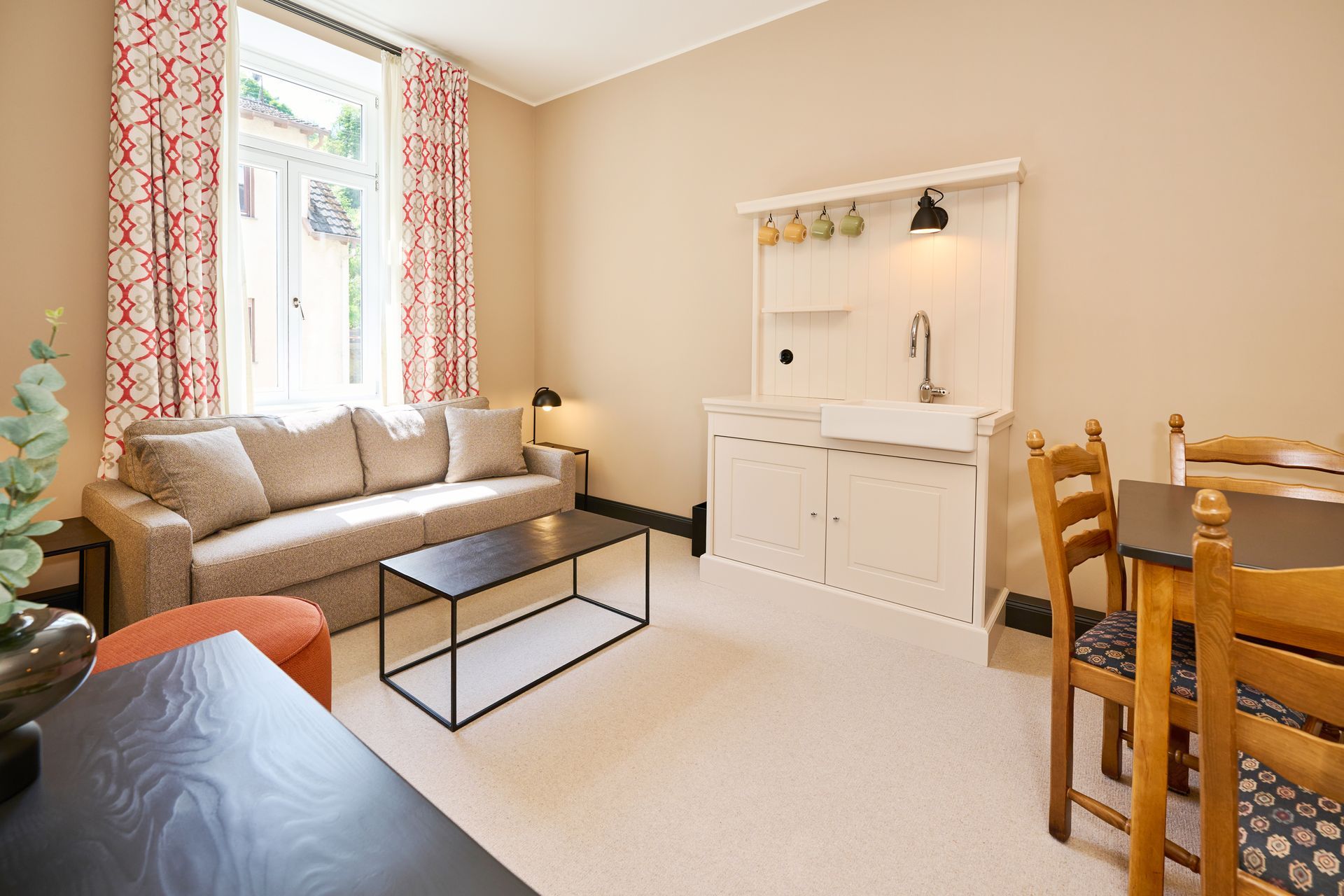 Ein Wohnzimmer mit beigefarbenem Sofa, schwarzem Couchtisch, kleiner Küchenzeile mit Spüle und einem Esstisch aus Holz.