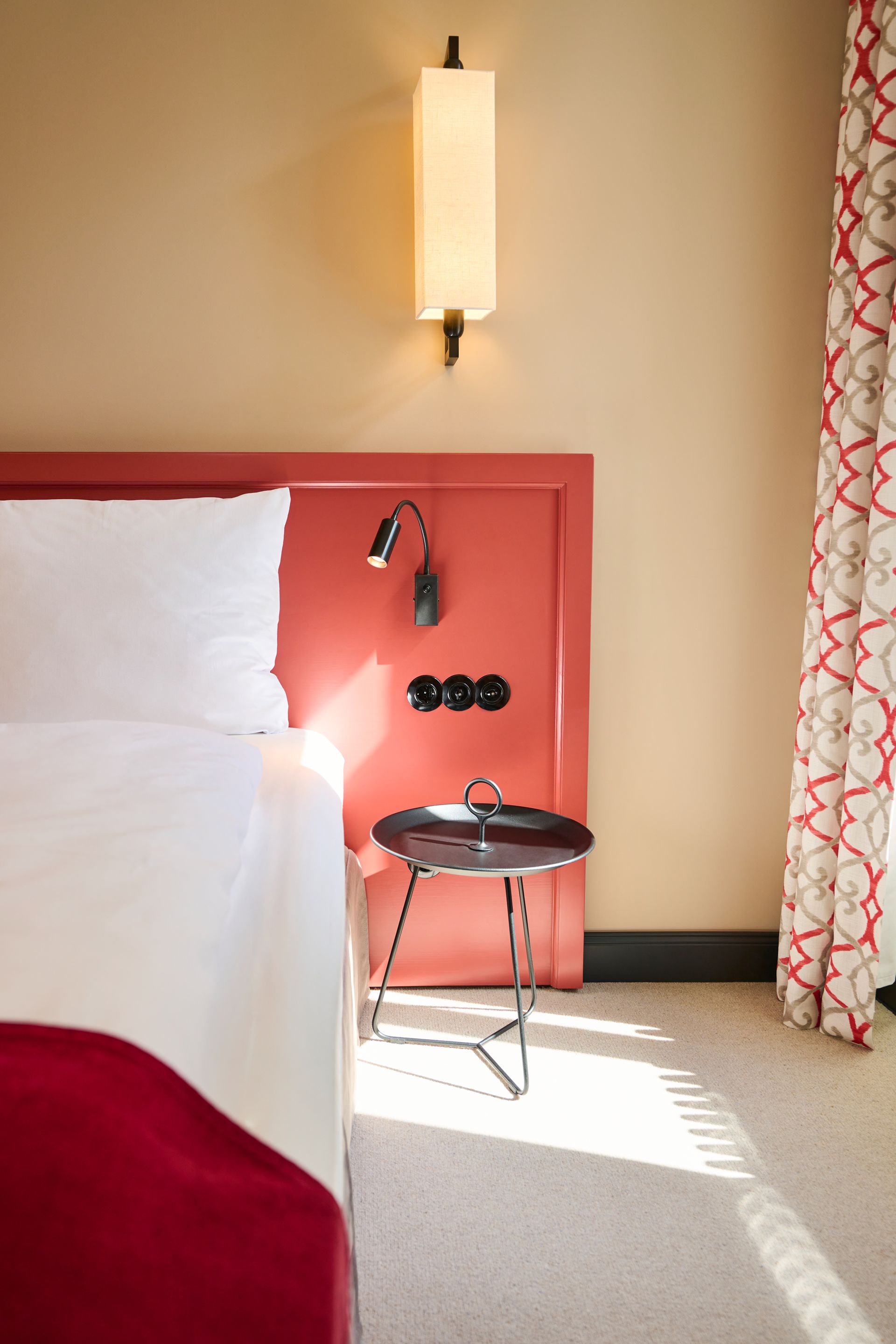 Eine moderne Schlafzimmerecke mit rotem Kopfteil, weiß bezogenem Bett, einem kleinen runden Tisch und Wandleuchten.