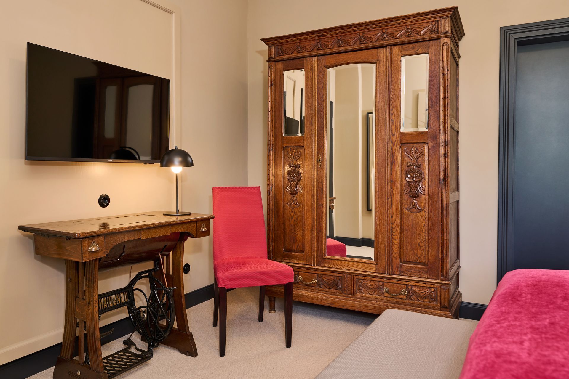 Ein Hotelzimmer mit einem Schreibtisch in Form einer alten Nähmaschine, einem leuchtend pinkfarbenen Stuhl, einem großen Holzschrank und einem an der Wand montierten Fernseher.