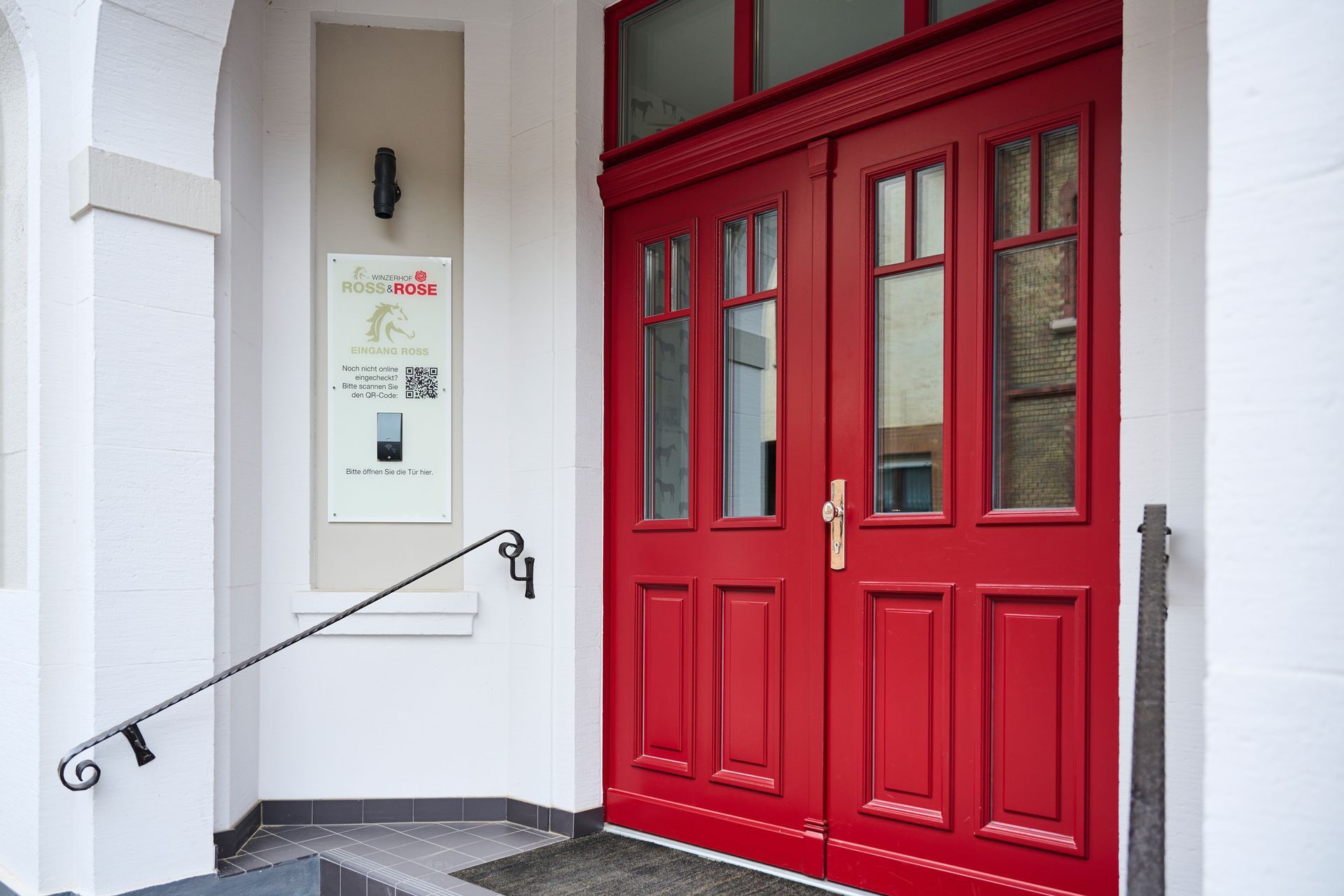 Ein Paar leuchtend rote Doppeltüren mit Glaseinsätzen am Eingang eines weißen Gebäudes, an dem ein Informationsschild angebracht ist.