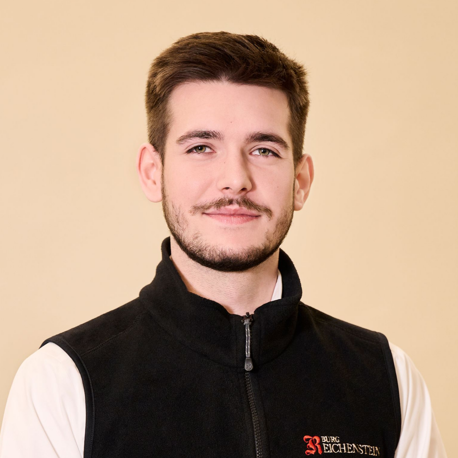 Eine Person mit kurzem braunem Haar und Bart, die eine schwarze Weste mit Reißverschluss über einem weißen Hemd trägt, vor beigem Hintergrund.