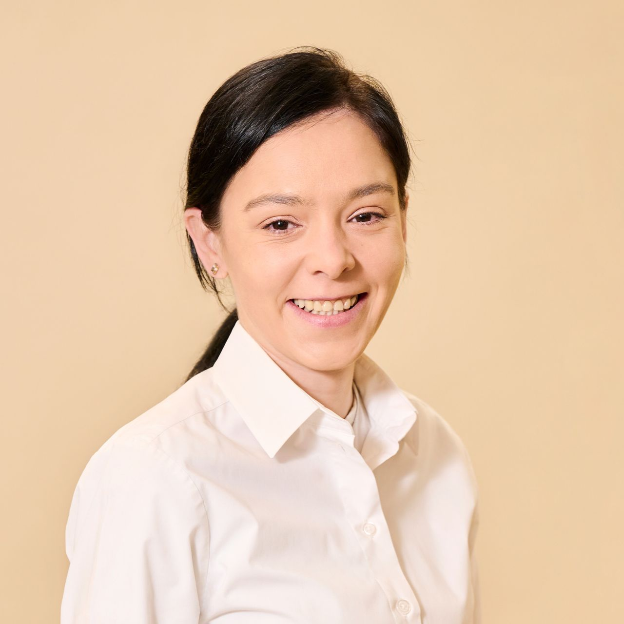 Eine lächelnde Person mit zurückgebundenem, dunklem Haar und einem weißen Hemd mit Knöpfen vor einem hellen, einfarbigen Hintergrund.