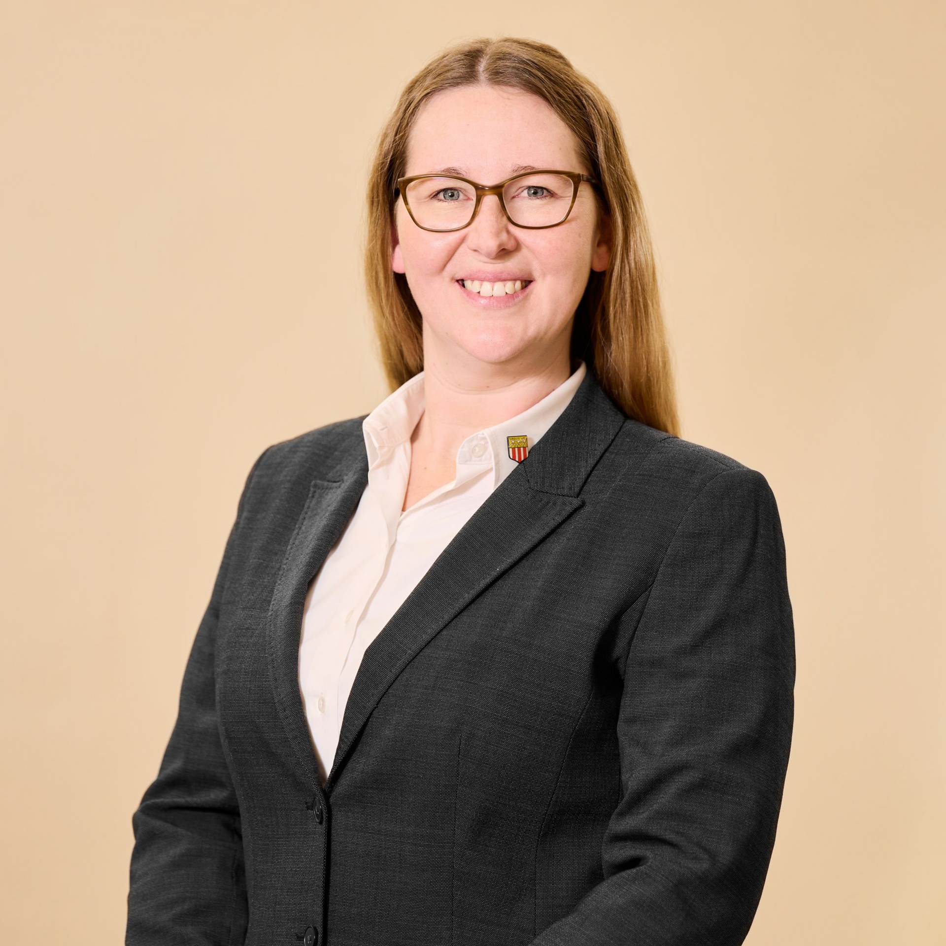 Eine Person in einem dunklen Blazer und einem weißen Hemd lächelt vor einem schlichten beigen Hintergrund.