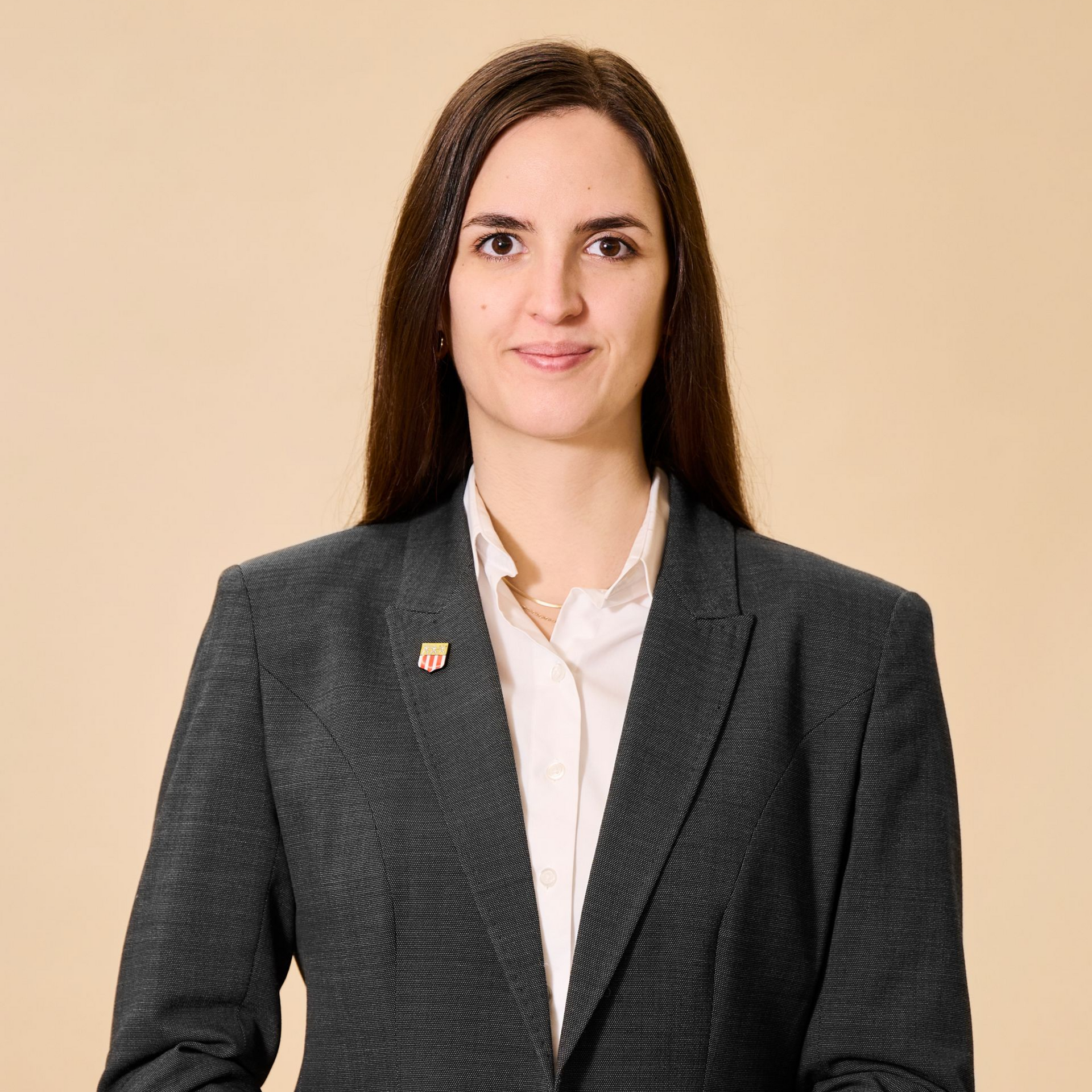 Professionelles Porträtfoto einer Person mit langen dunklen Haaren, die ein weißes Hemd und einen dunklen Blazer vor einem beigen Hintergrund trägt.