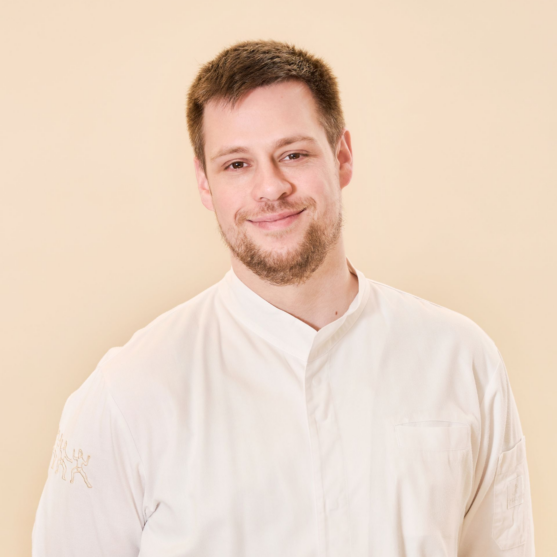 Eine Person mit kurzem Bart, die eine weiße Kochjacke trägt und vor einem schlichten, hellen Hintergrund lächelt.