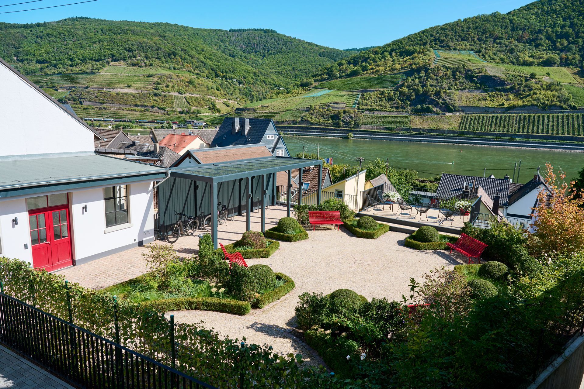 Eine sonnige Terrasse mit roten Bänken und einer Pergola bietet Blick auf ein Flusstal mit grünen, terrassenförmig angelegten Hügeln und einem kleinen Dorf.