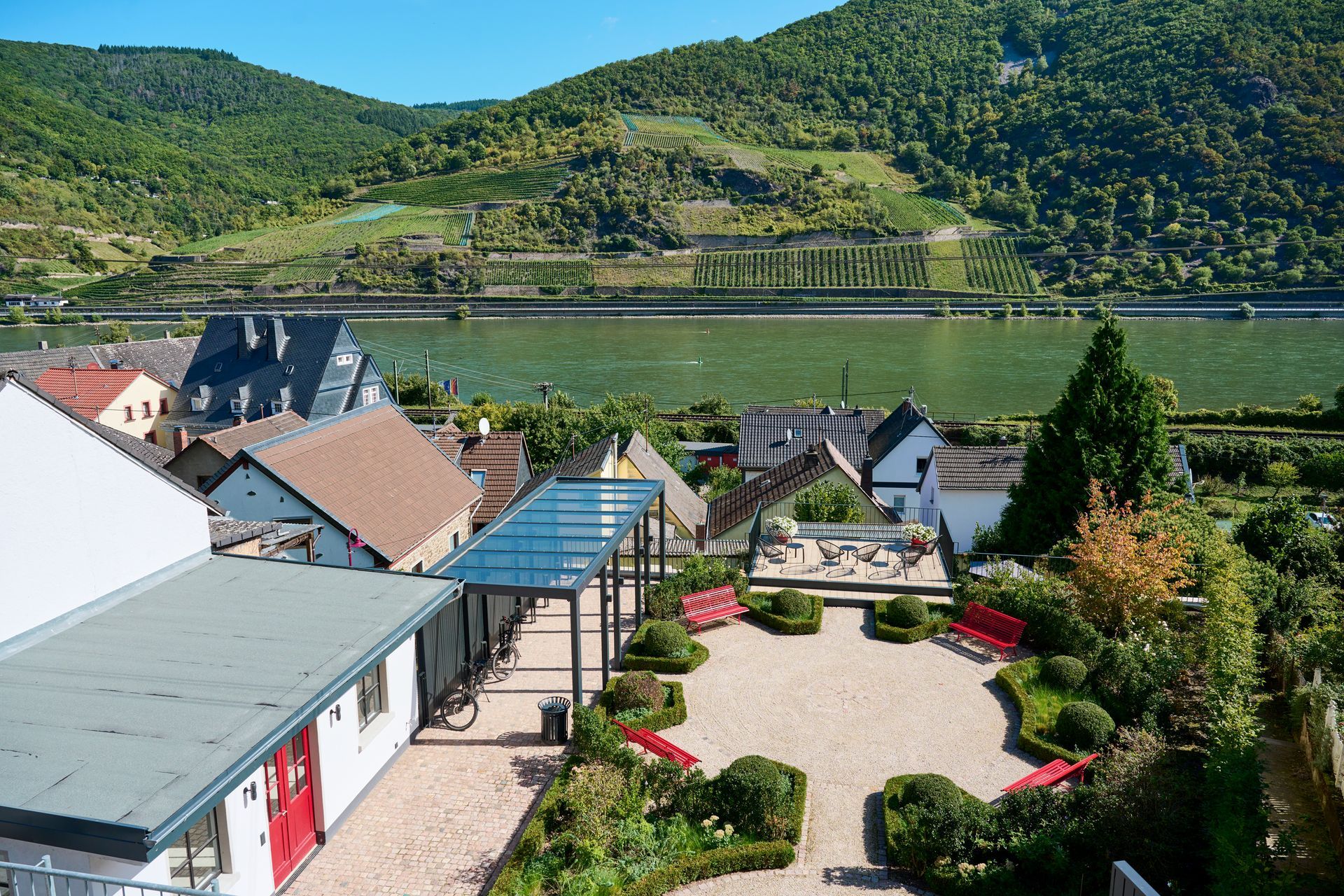 Ein mit Kies bedeckter Innenhof mit roten Bänken, einer Glaspergola und nahegelegenen Häusern, eingebettet in die Kulisse eines Flusses und bewaldeter Hügel.