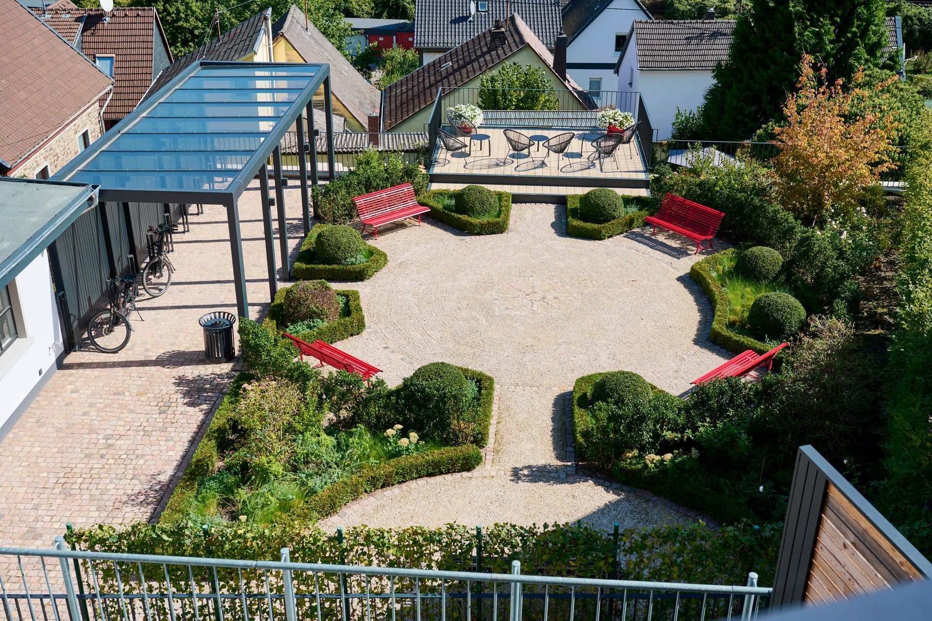 Ein erhöhter Blick auf einen landschaftlich gestalteten Innenhof mit Kieswegen, akkurat geschnittenen Hecken, mehreren roten Bänken und einem gläsernen Pavillon.