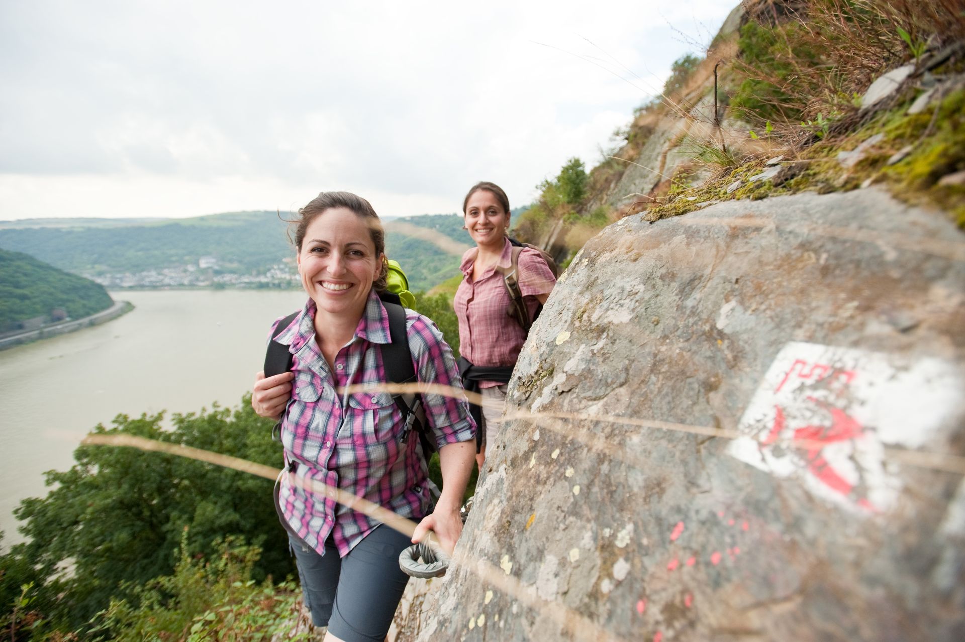 Zwei Personen wandern auf einem schmalen, felsigen Pfad an einer Klippe entlang, der einen breiten Fluss überblickt, und nutzen dabei ein Sicherheitsseil zur Unterstützung.