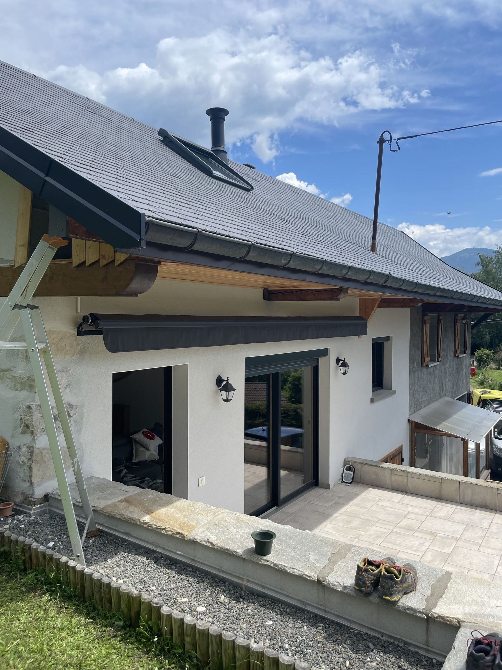 Extérieur de la maison avec murs en stuc blanc, toit de tuiles grises et portes-fenêtres sous un ciel bleu.