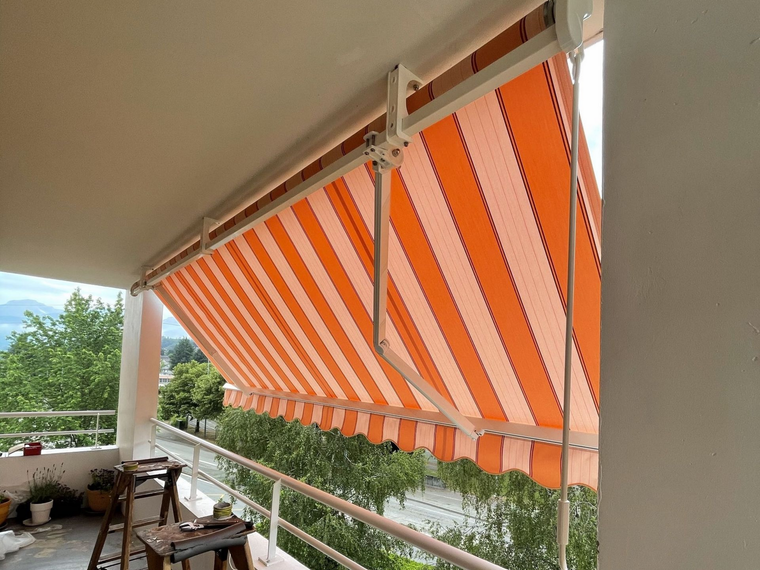 Un auvent à rayures orange protège une véranda avec rambarde, chaises et arbres à l'extérieur.