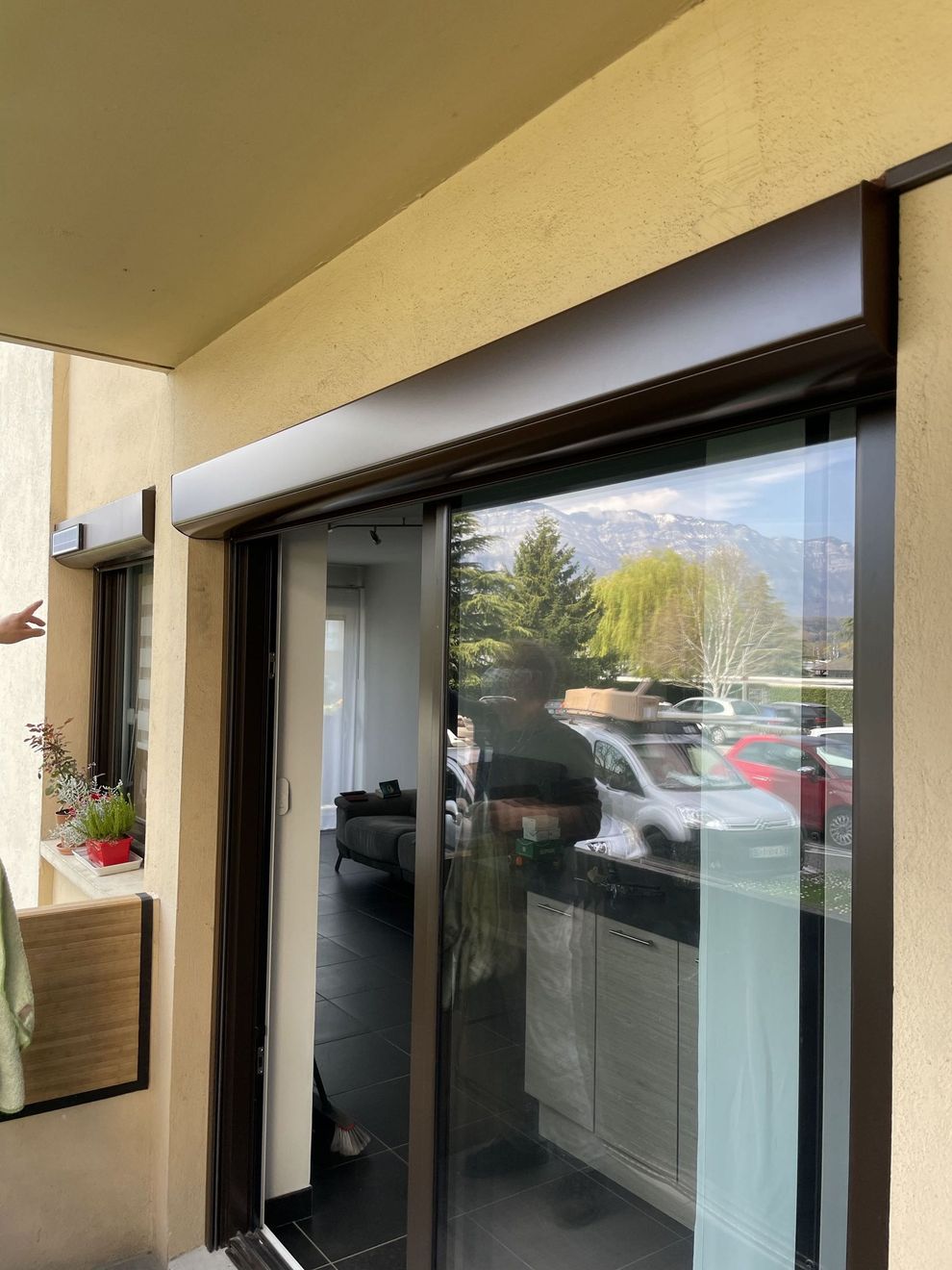 Balcon d'appartement avec porte coulissante en verre, mur ocre et reflets de voitures garées et de montagnes.