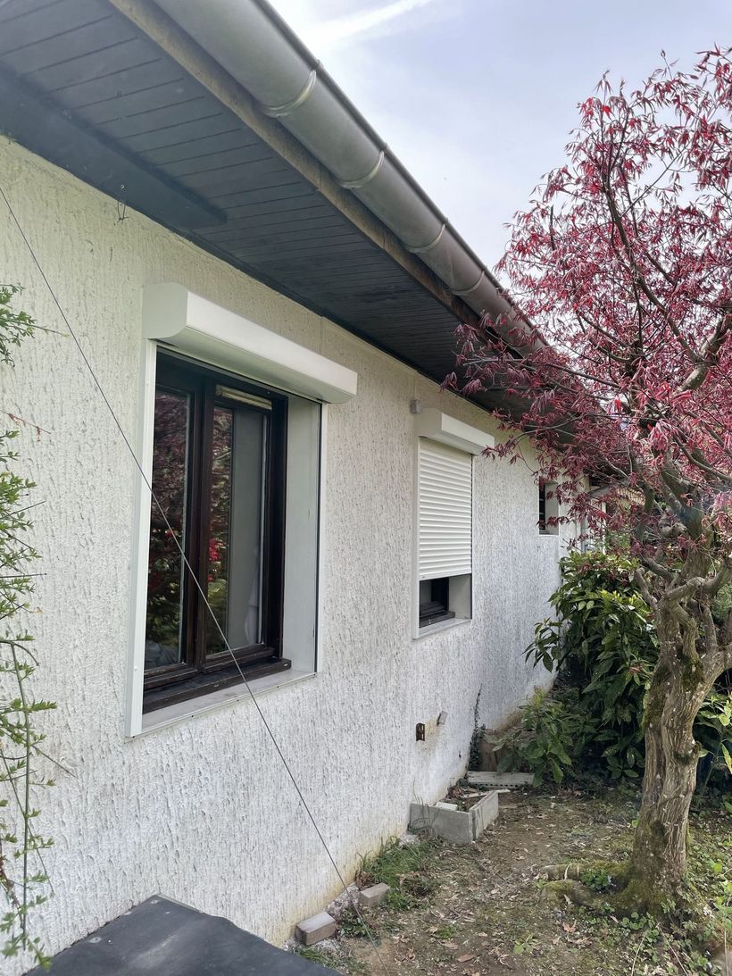 Maison blanche avec fenêtre à encadrement noir, petite aération et un arbre à feuilles rouges près du mur.