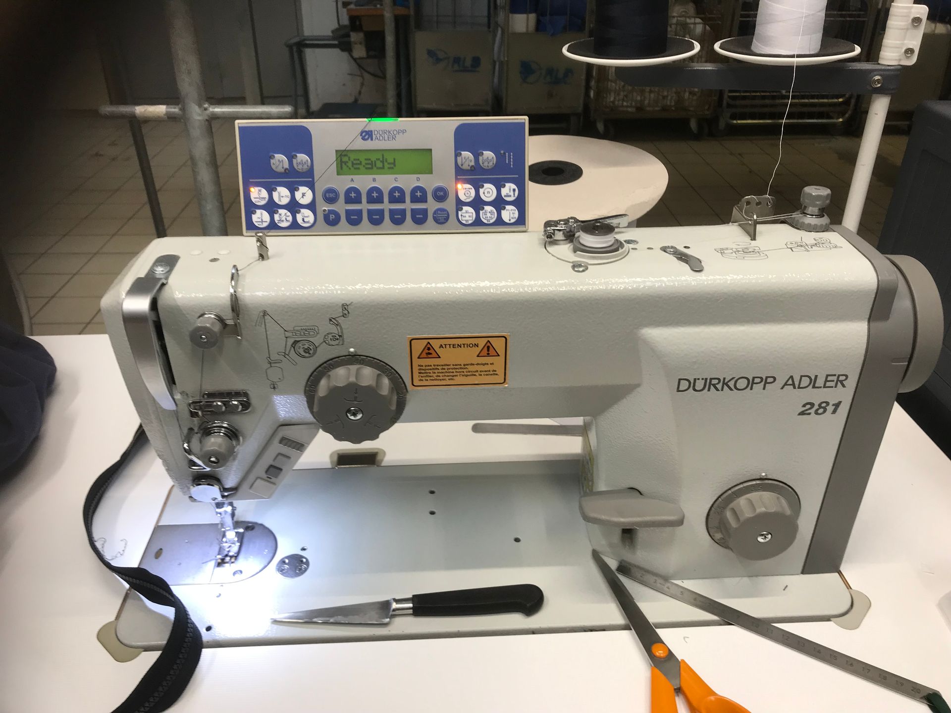Une machine à coudre industrielle grise Dürkopp Adler 281 sur une table blanche, avec des ciseaux et un découseur à proximité.