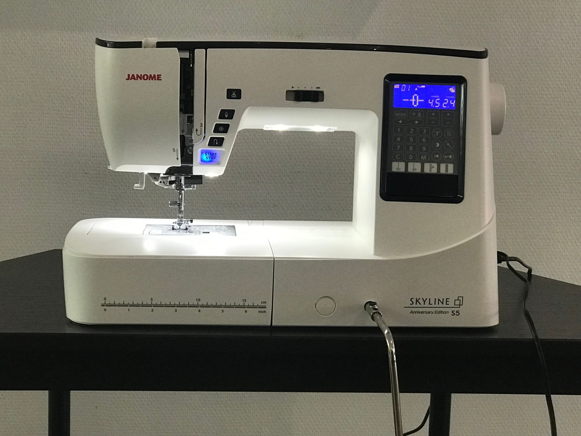 Une machine à coudre informatisée Janome blanche est posée sur une table noire, sa lampe de travail allumée et son écran d'affichage actif.