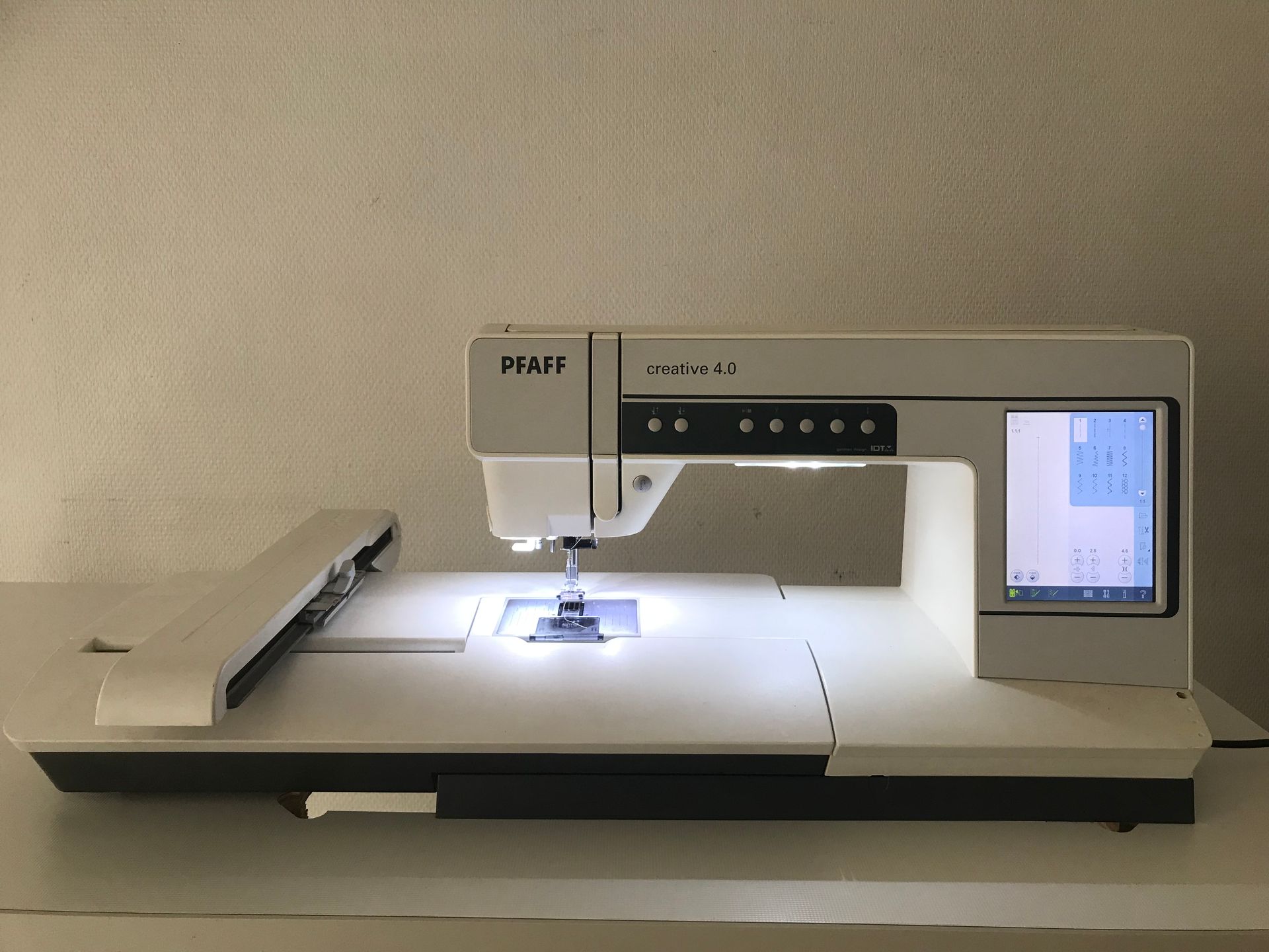 Une machine à broder Pfaff blanche est posée sur une surface plane, avec un espace de travail éclairé et un écran tactile numérique.