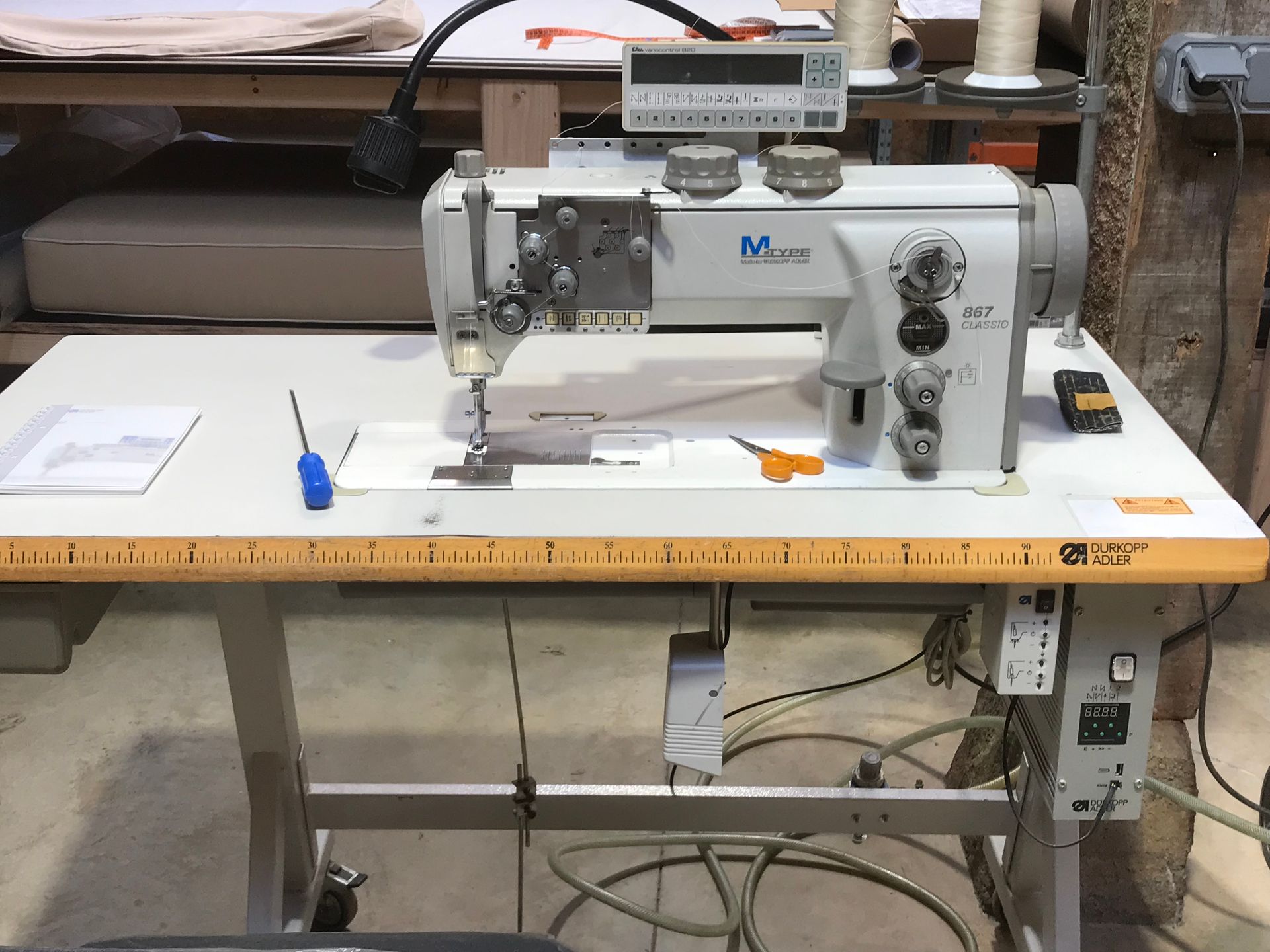 Une machine à coudre industrielle posée sur une table claire dans un atelier, équipée d'un écran numérique, d'outils et d'un bloc d'alimentation.