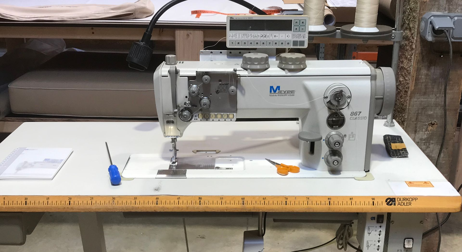 Une machine à coudre industrielle blanche, dotée d'un affichage numérique, est posée sur une table de travail, avec un tournevis et des ciseaux.