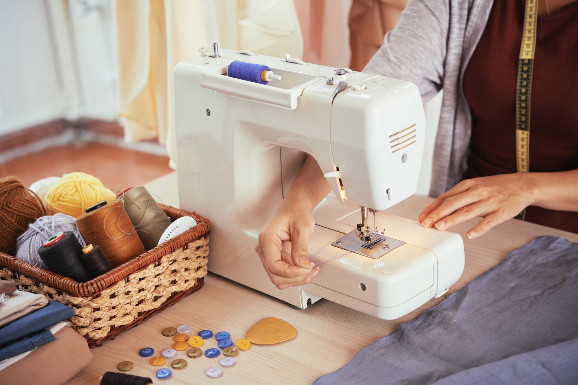 Une personne utilise une machine à coudre blanche pour coudre du tissu, avec un panier de bobines de fil et des boutons éparpillés sur la table.