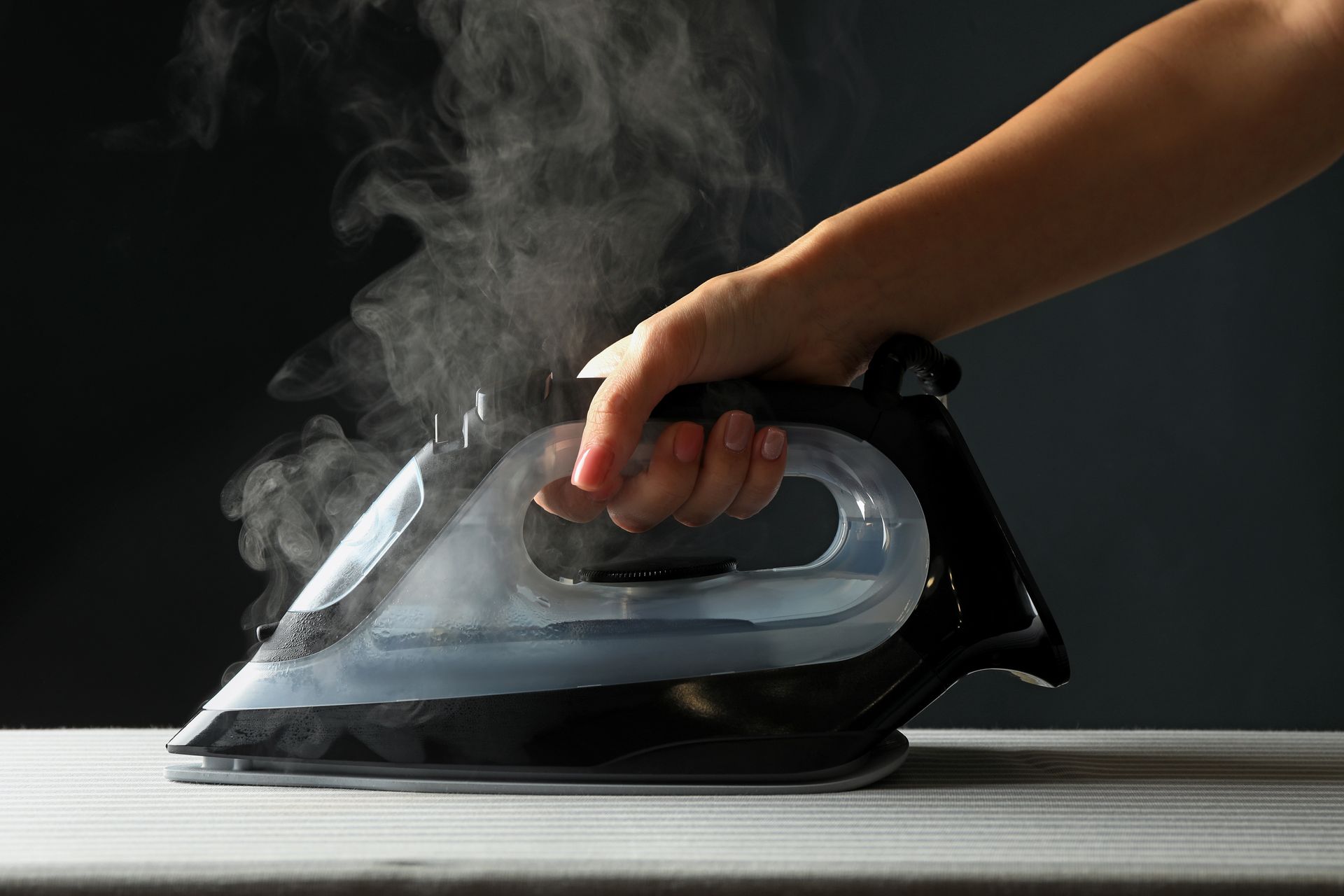 Une main tient un fer à repasser à vapeur noir au-dessus d'une surface plane, de la vapeur s'en échappant sur un fond sombre.