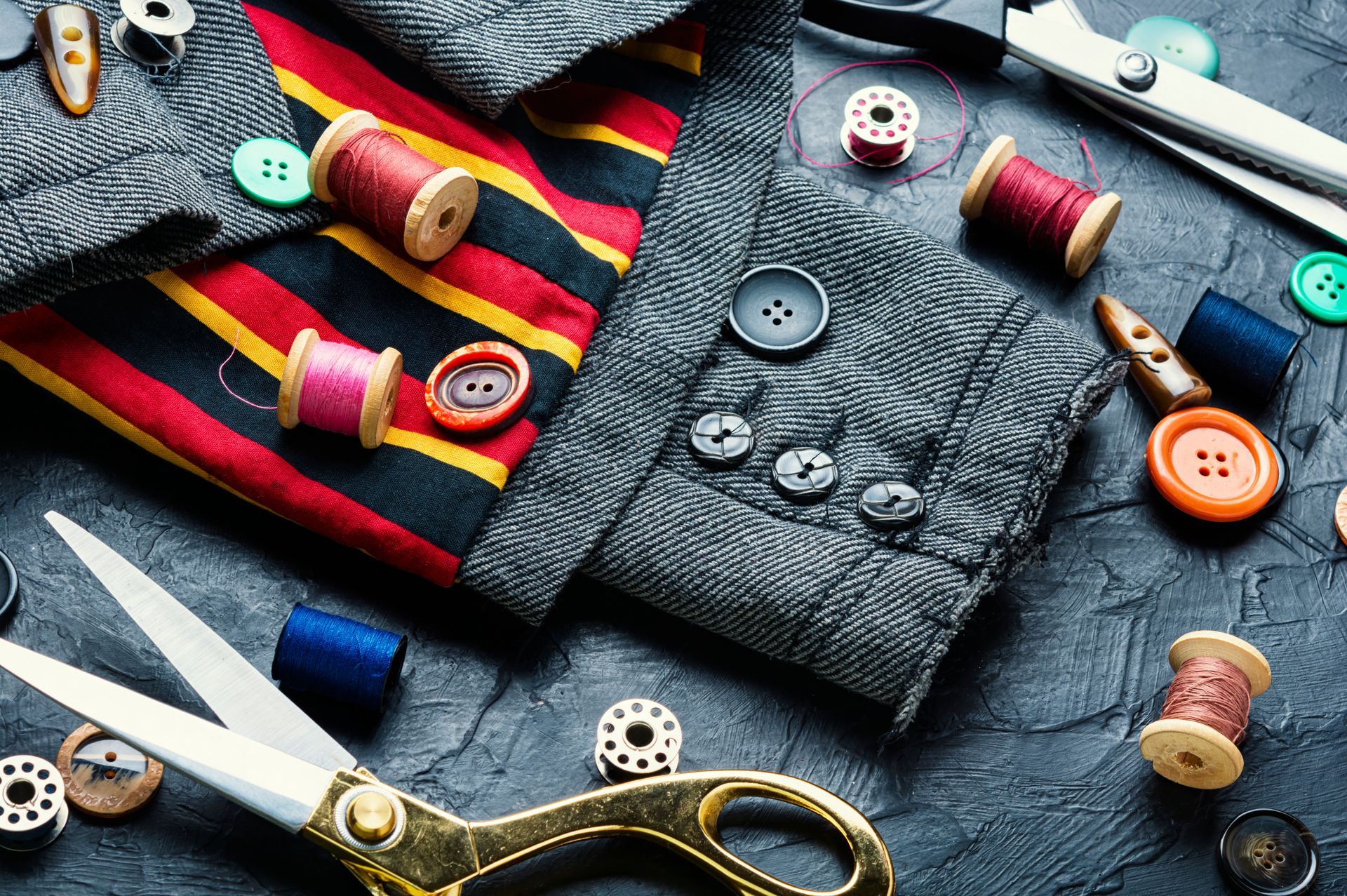 Fournitures de couture, notamment des bobines de fil, des boutons colorés et des ciseaux, disposées sur une surface sombre et texturée.
