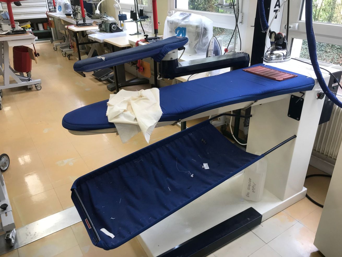 Poste de repassage industriel avec une planche rembourrée bleue, un bac de récupération des lingettes et un fer à repasser dans un atelier de couture bien éclairé.