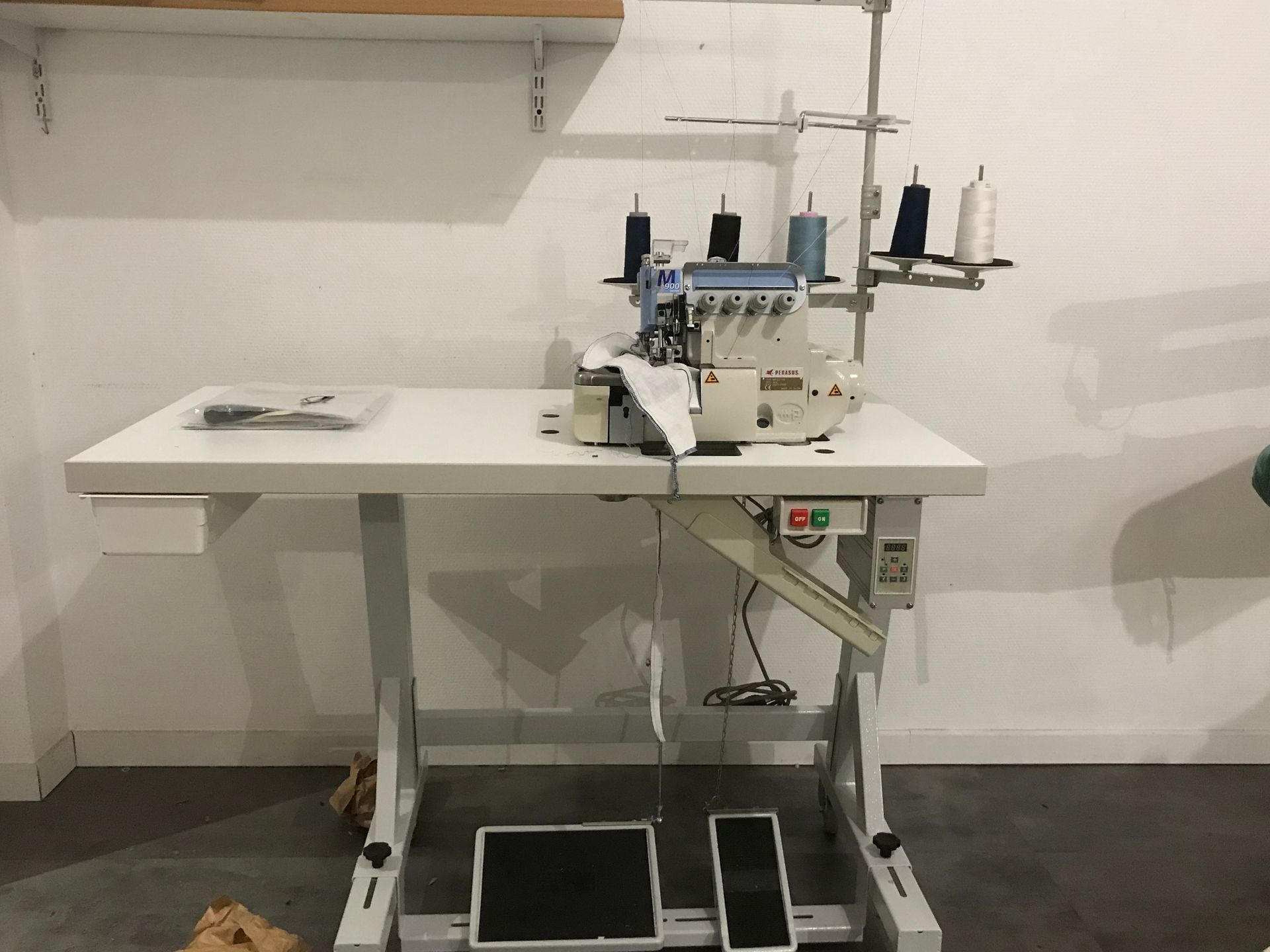 Une surjeteuse industrielle posée sur une table blanche avec des cônes de fil, devant un mur blanc uni.