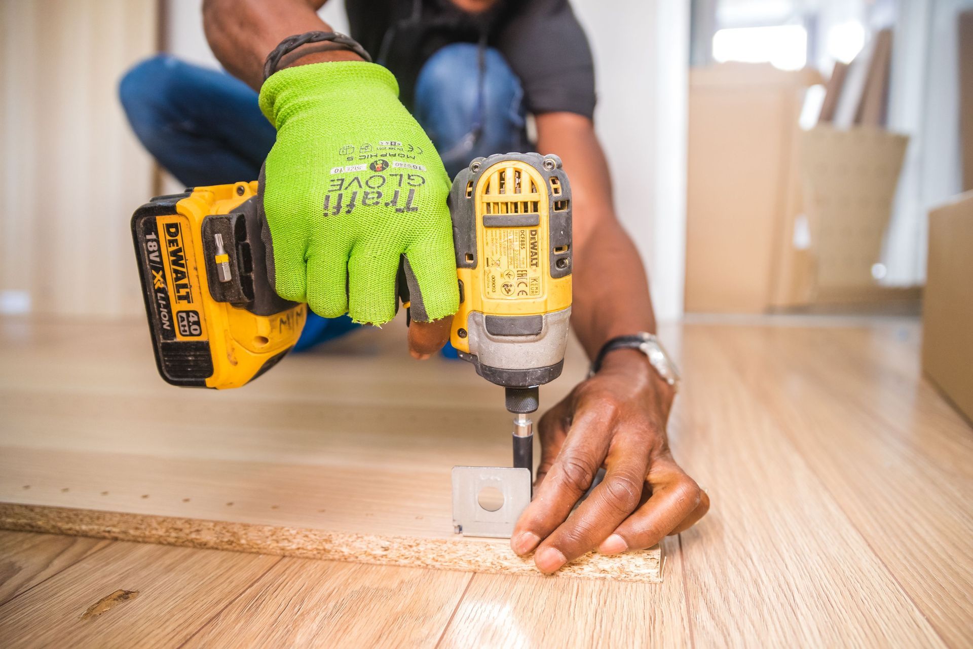 Personne utilisant une perceuse jaune pour visser une vis dans le sol, avec un gant vert, sur un plancher en bois
