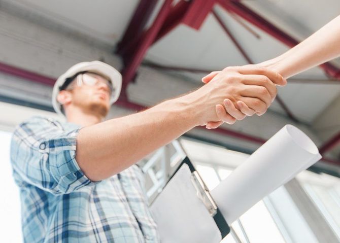 Deux personnes se serrent la main dans un espace de travail industriel lumineux, l'une d'elles portant un casque et un bloc-notes.