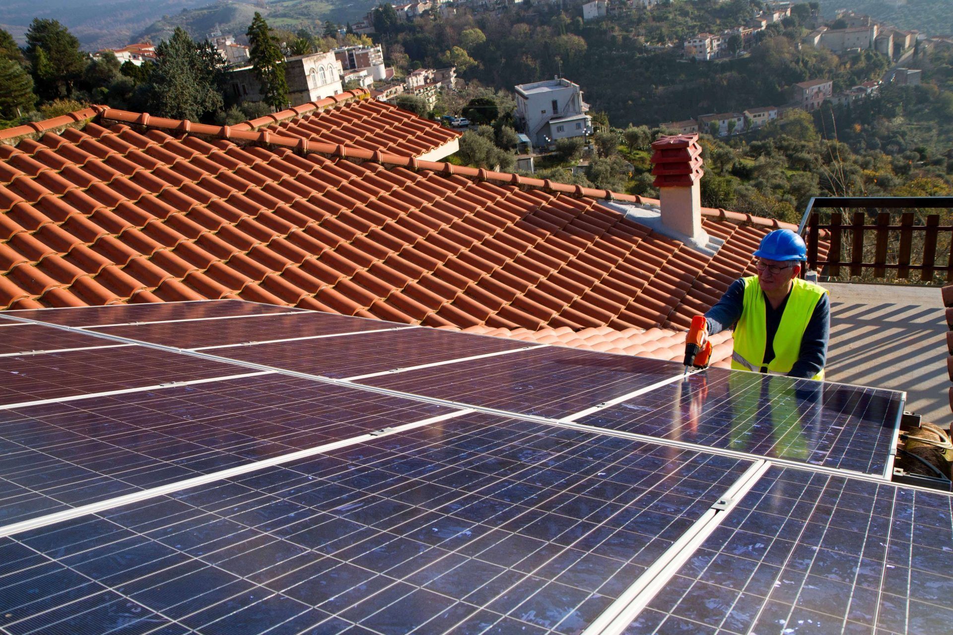 Ouvrier nettoyant des panneaux solaires sur un toit de tuiles, avec des collines en arrière-plan.