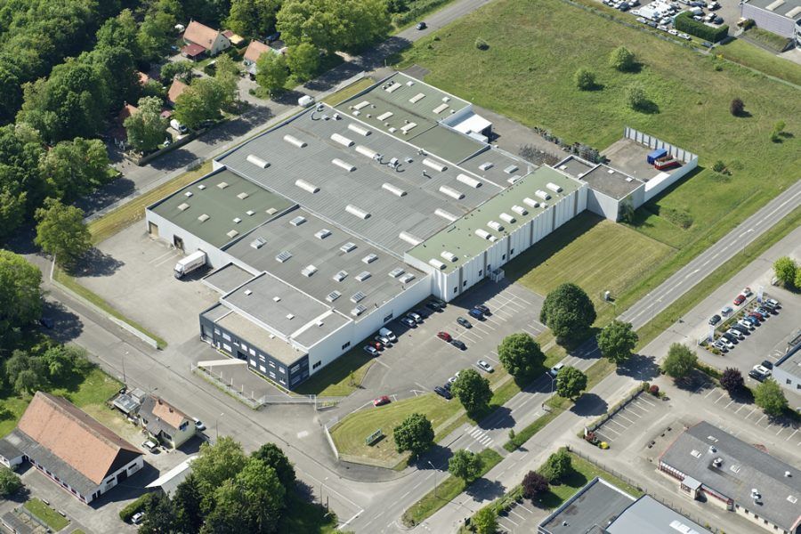 Vue aérienne d'un grand bâtiment industriel gris avec des parkings, des arbres et des routes à proximité.