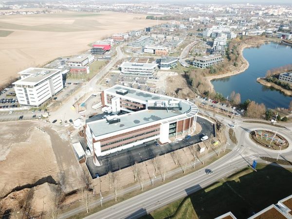 Vue aérienne d'un bâtiment moderne en construction ou en cours d'aménagement, situé au bord d'un lac, de routes et d'un terrain vague.