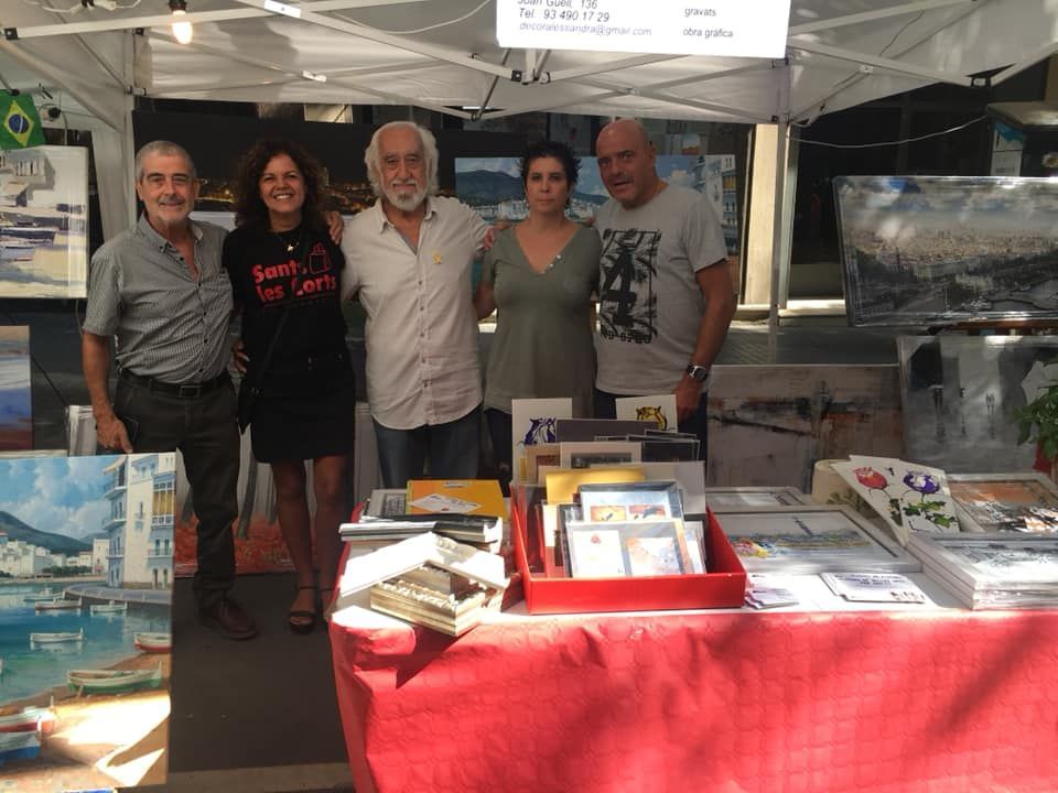 Cinco personas posan juntas detrás de una mesa cubierta de rojo donde se exhiben láminas artísticas en una feria de artesanía al aire libre.