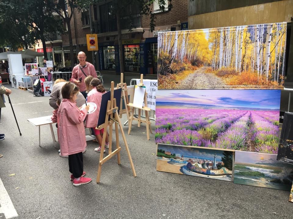 Niños pintan en caballetes en una calle de la ciudad, al aire libre, junto a grandes lienzos impresos con paisajes.