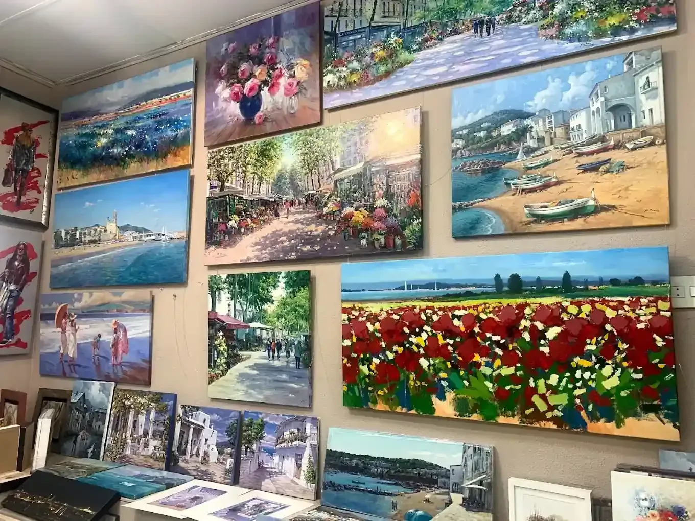 Una pared de exposición con varios cuadros al óleo enmarcados de paisajes, flores y escenas marinas en una galería o tienda de arte.